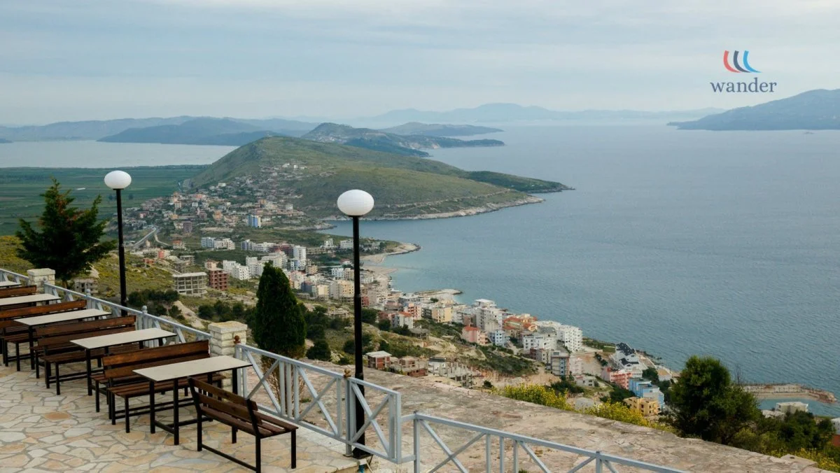 A scenic view of a coastal town with colorful buildings, a large body of water, and green hills in the background, taken from an outdoor terrace with benches and lamp posts.