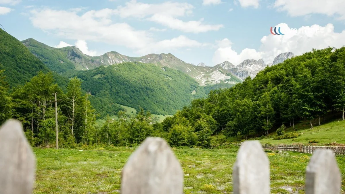 Scenic view of green mountains and forested hills under a partly cloudy sky, with a wooden fence in the foreground.