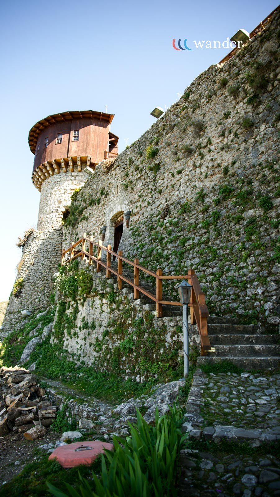 A stone castle wall with a wooden staircase leading to a door, a turret with a wooden structure on top, surrounded by greenery and a clear blue sky.