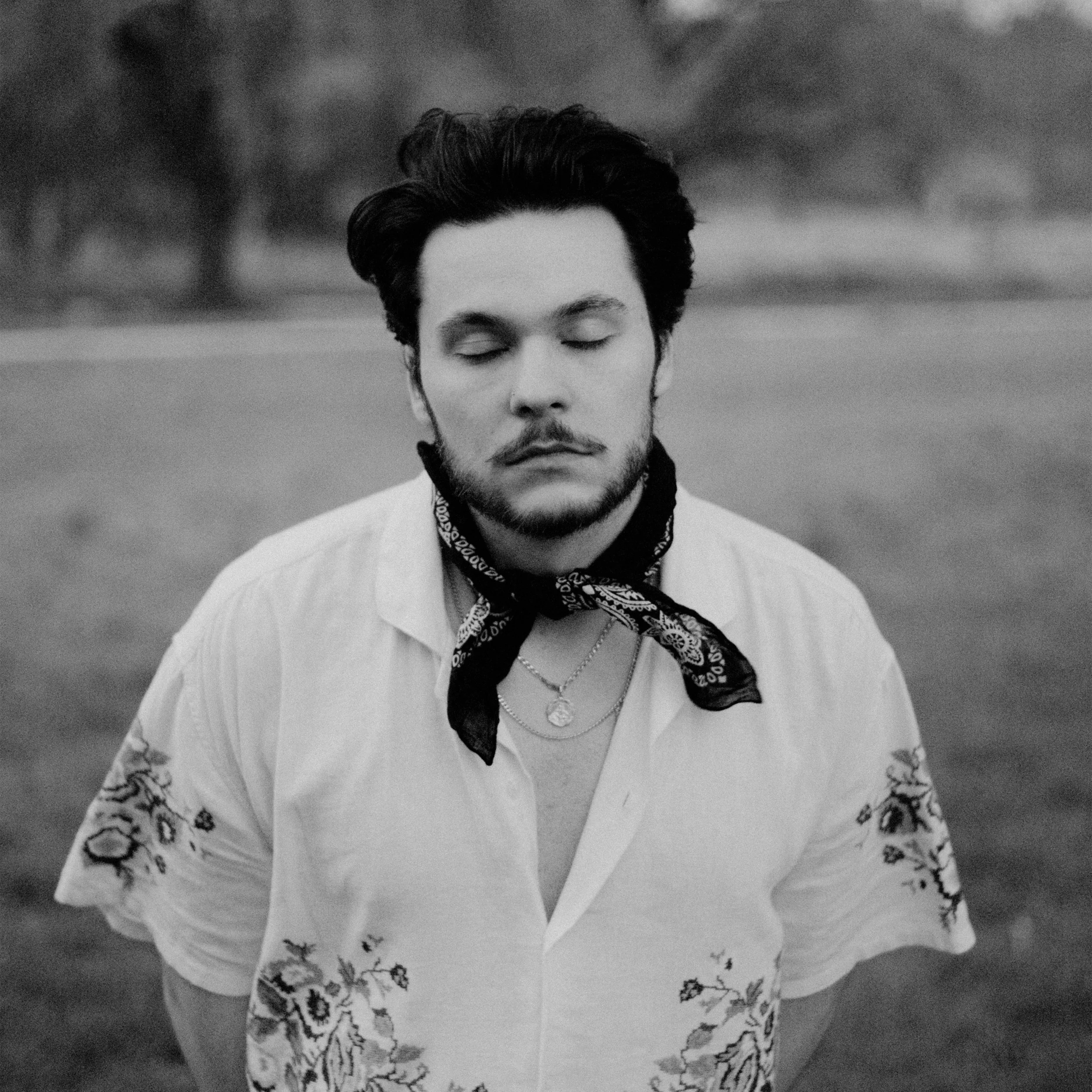 Black and white photo of a young man with closed eyes, wearing a embroidered short-sleeve shirt, a bandana around his neck, and layered necklaces, outdoors with blurred trees in the background.