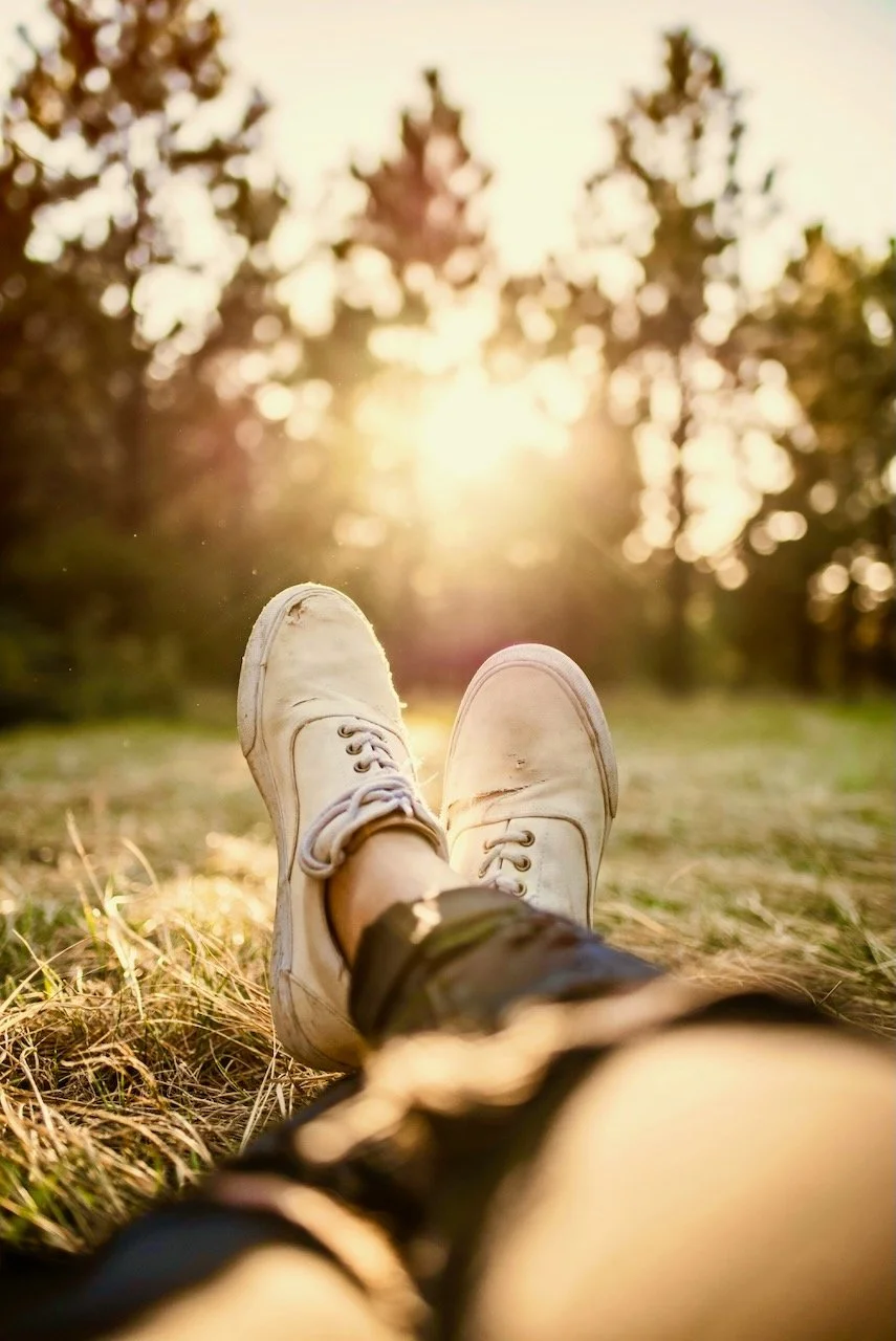 View of a relaxed, peaceful person lying on the grass with legs stretched out, wearing white sneakers and black pants, in a park with trees and sunlight in the background.