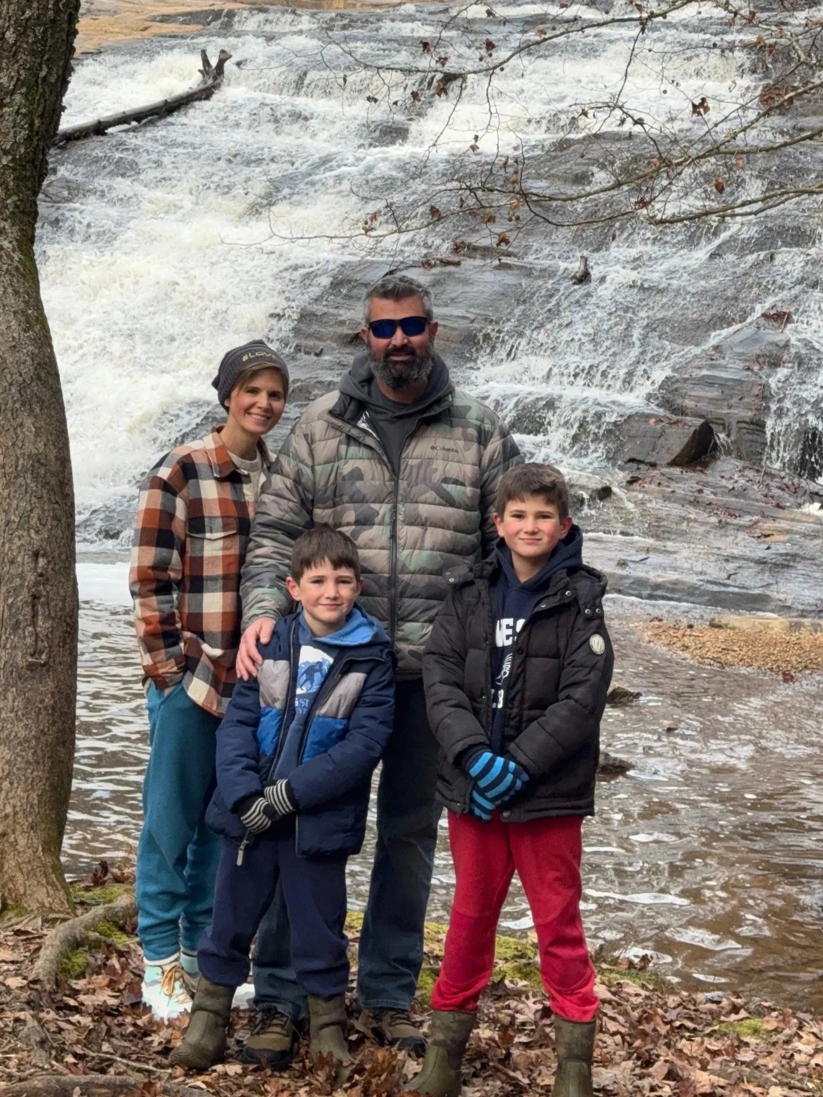 A family of four stands in front of a waterfall with rocky terrain and fallen leaves on the ground. The family includes two children, a woman, and a man, all dressed warmly for outdoor fall weather.