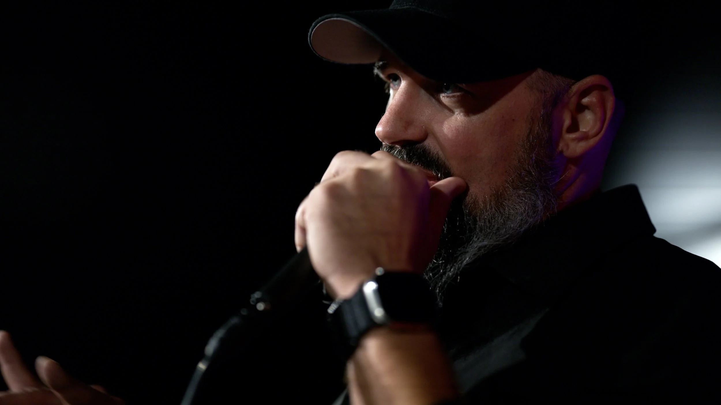 Close-up of a man wearing a black baseball cap and black shirt, holding a microphone near his mouth, in a dark setting with a faint light illuminating his face.