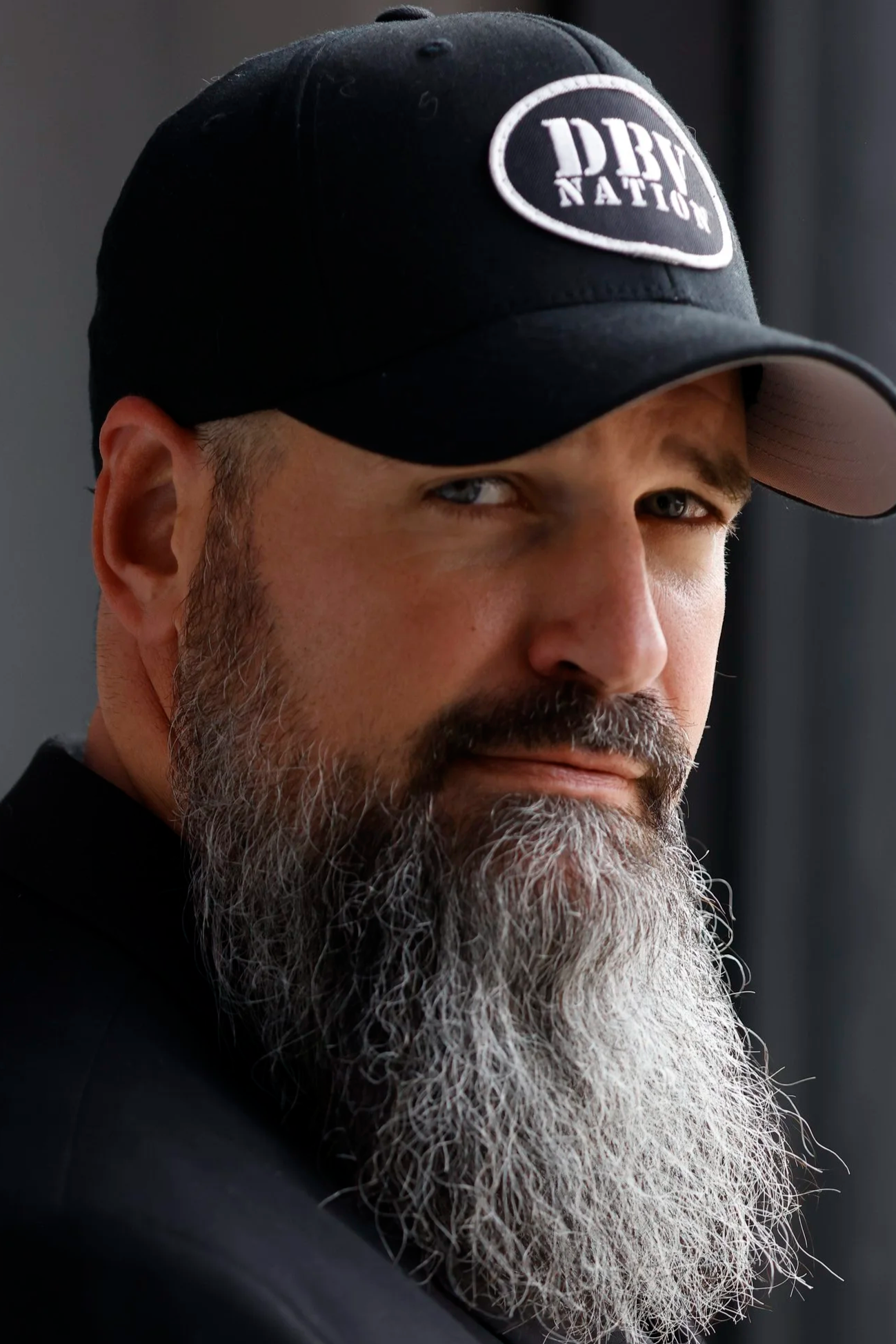 A middle-aged man with a gray beard wearing a black cap with 'DBD Nation' embroidered on it, looking towards the camera with a serious expression.