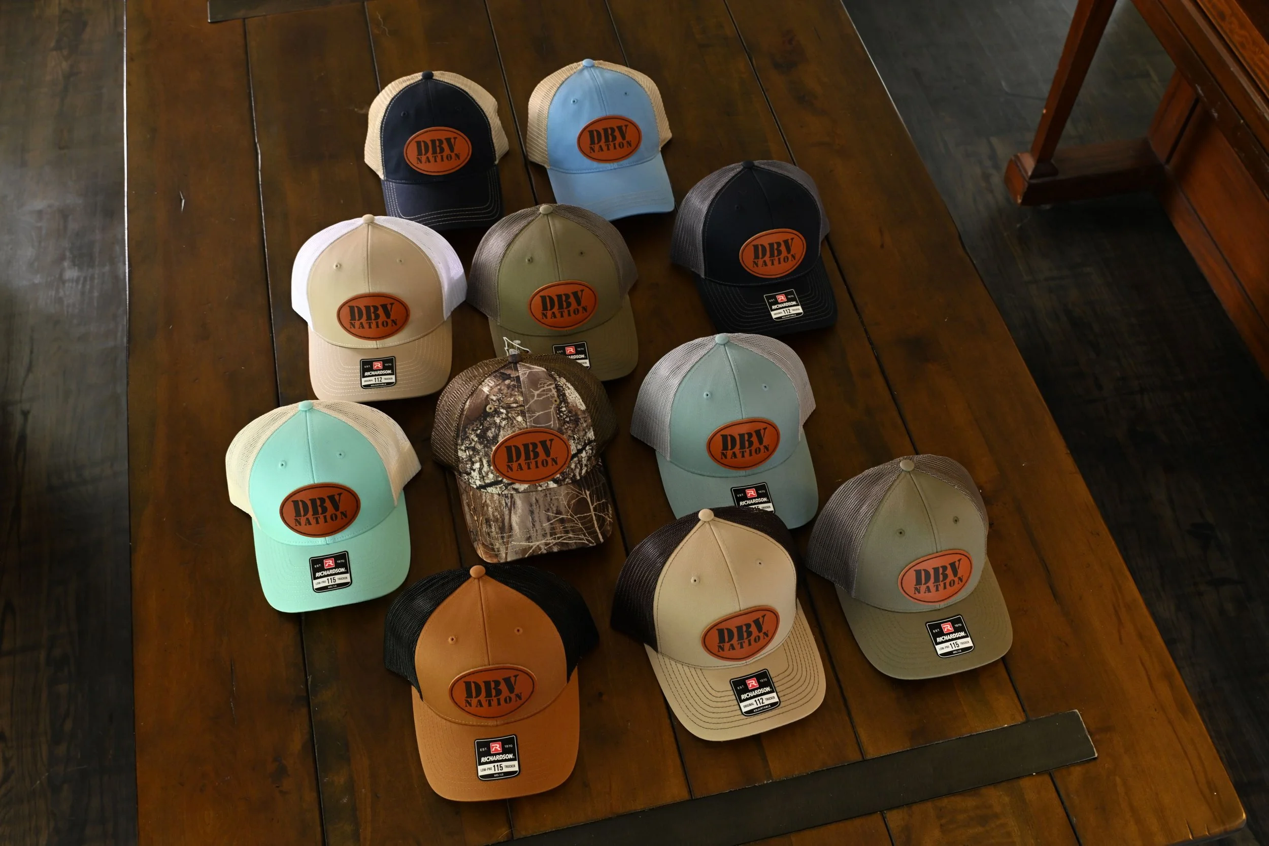A wooden table displaying 13 baseball caps with various colors and patterns, all featuring a large orange oval patch with black text reading 'DBV NATION' on the front.