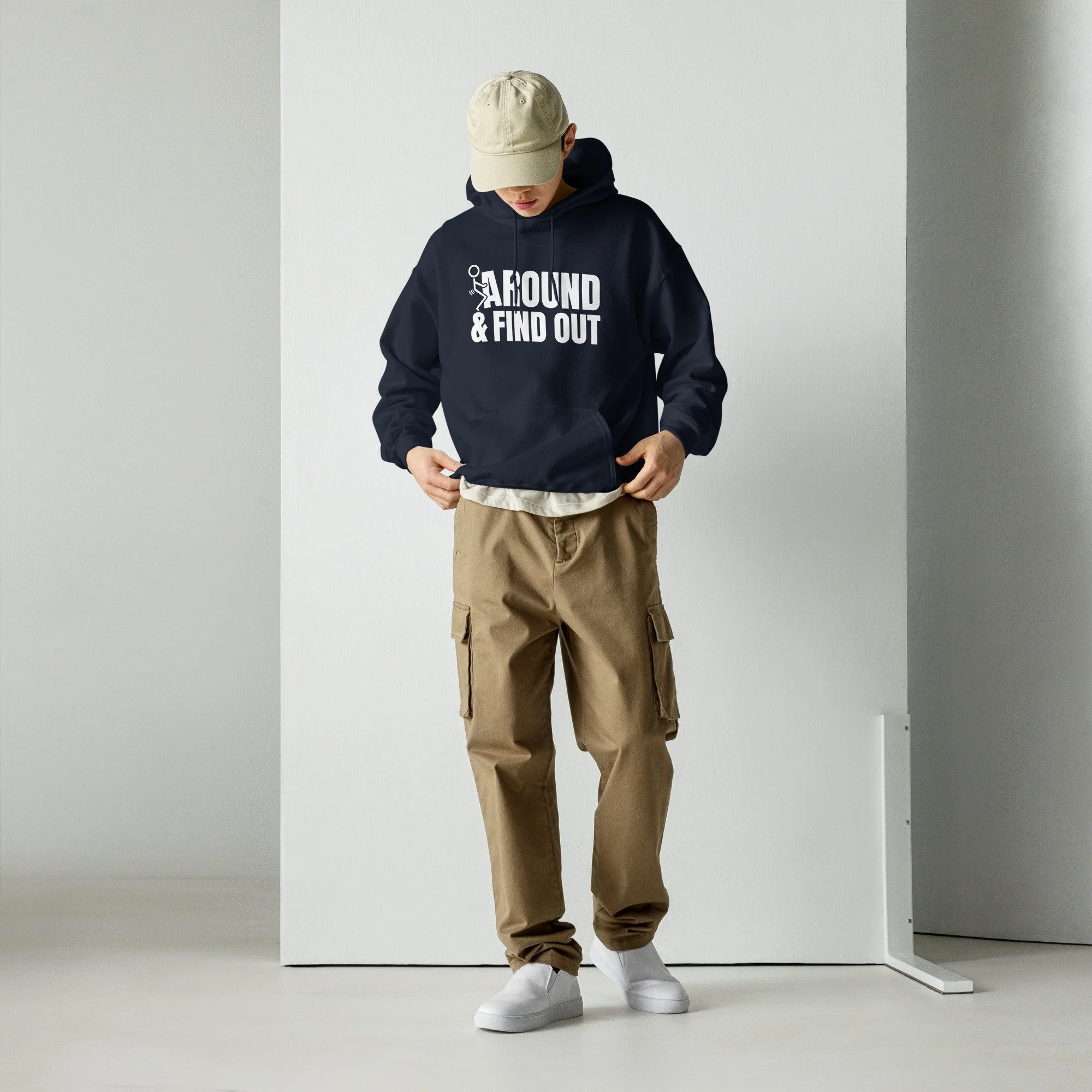 Young person wearing a beige cap, black hoodie with white text saying 'AROUND & FIND OUT', beige cargo pants, and white sneakers, standing against a plain background.