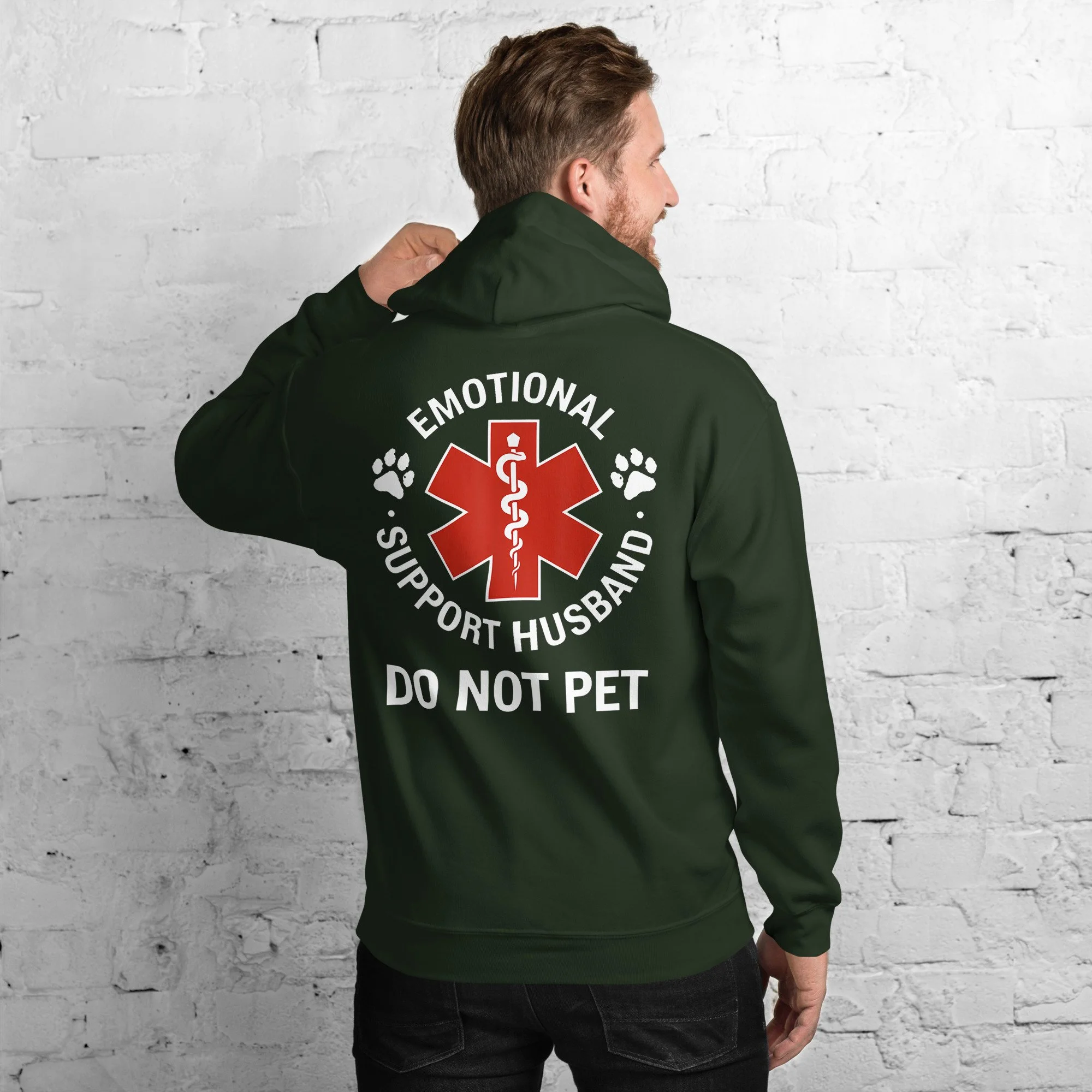 Back of a man wearing a green hoodie with a graphic design that says "Emotional Support Husband," featuring a red medical symbol and paw prints, advising not to pet the dog.