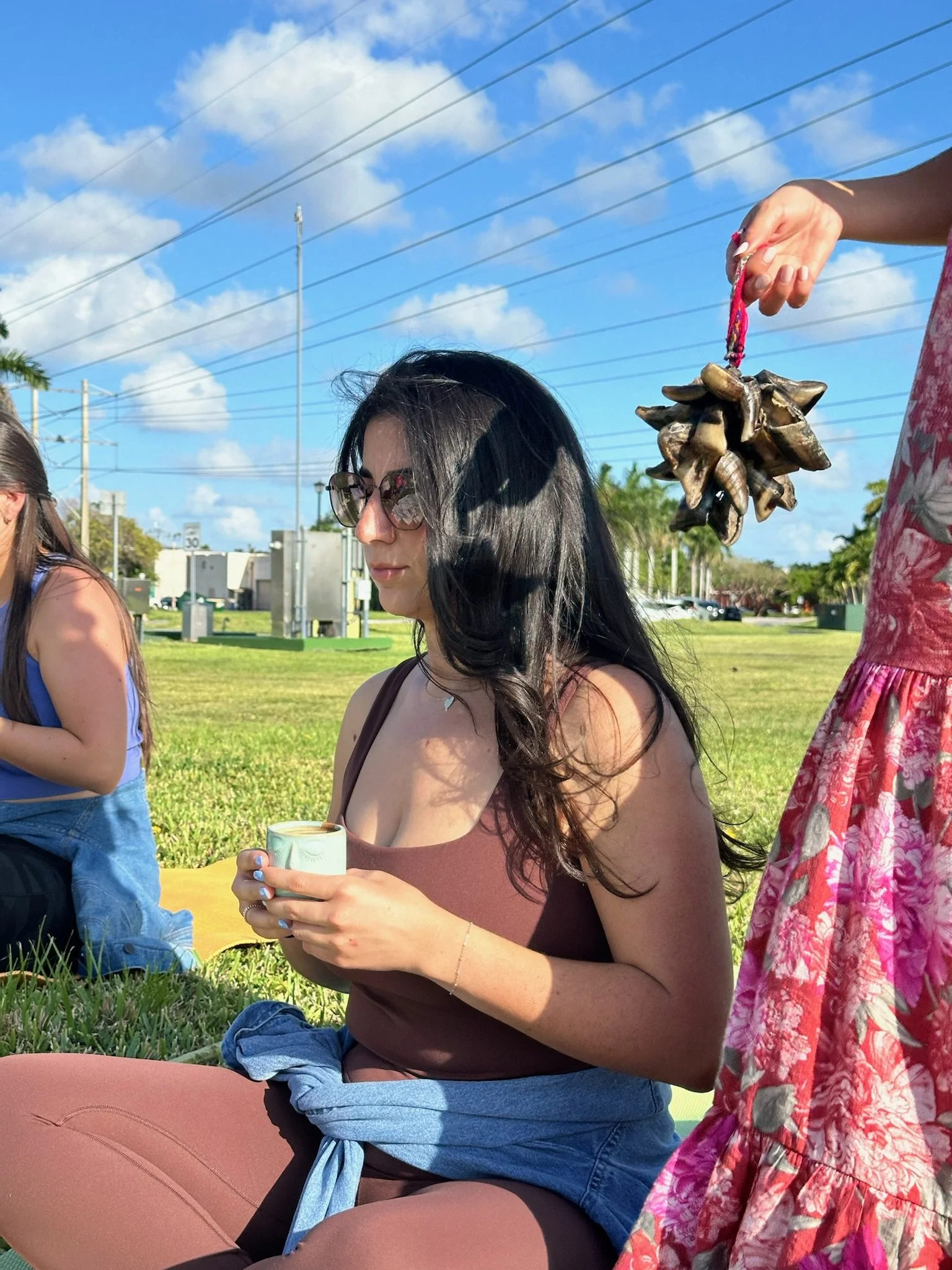 A woman lying on the grass with flowers on her chest, with her eyes closed and a serene expression, during sunset. Another person is sitting nearby with part of their arm and leg visible.