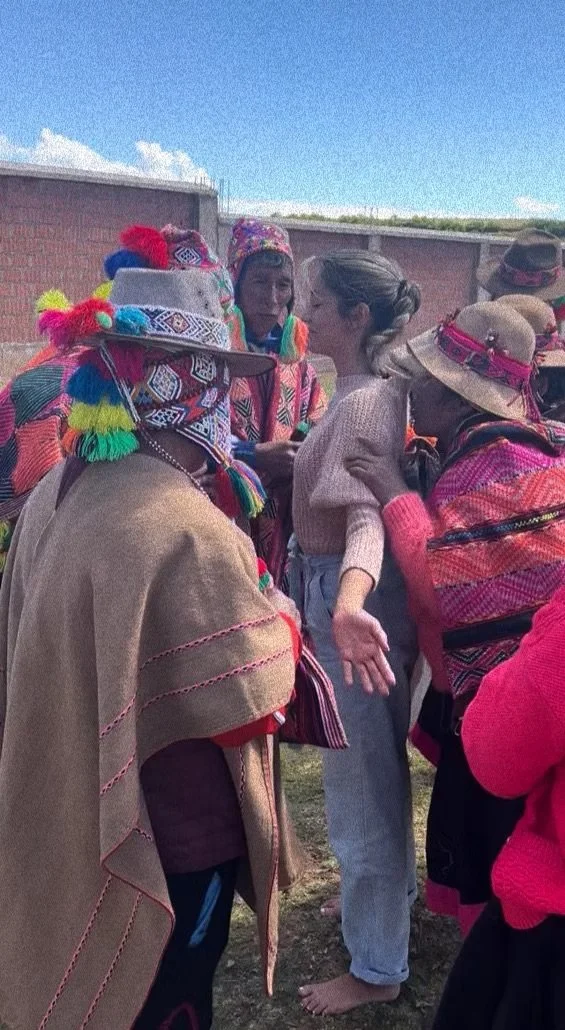 A woman is surrounded by members of an indigenous community dressed in colorful traditional clothing and hats, engaging in a close interaction outdoors on a sunny day.