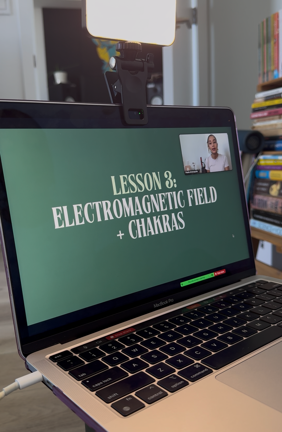 Laptop displaying a presentation slide titled 'Lesson 3: Electromagnetic Field + Chakras' during a virtual lesson. A small video window shows a woman speaking, and a desk lamp is positioned behind the laptop.