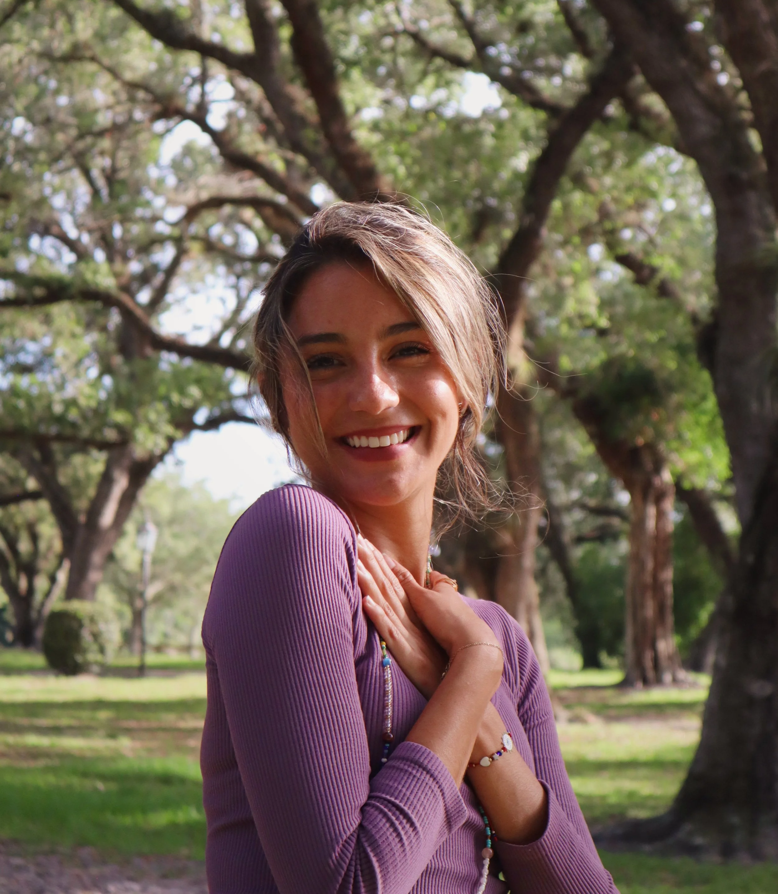Paola Amezquita with short hair, wearing a purple top, standing outdoors in a park with large trees and green grass.