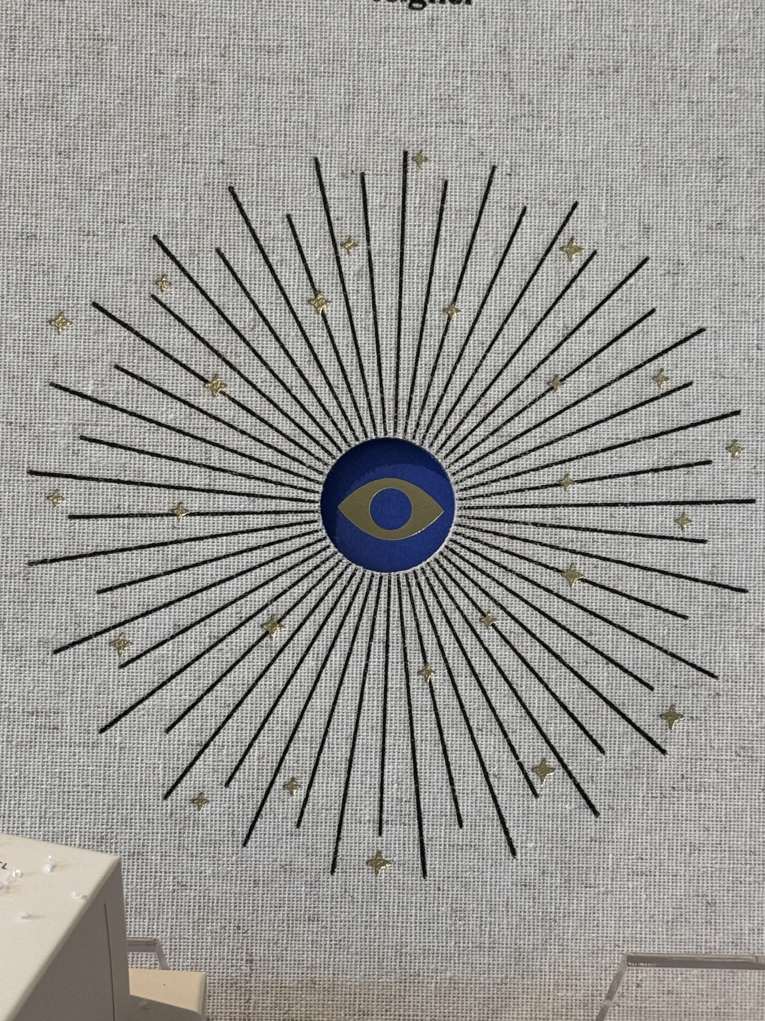 Decorative wall art with an eye symbol at the center, surrounded by radiating black lines and small gold stars on a textured gray background.