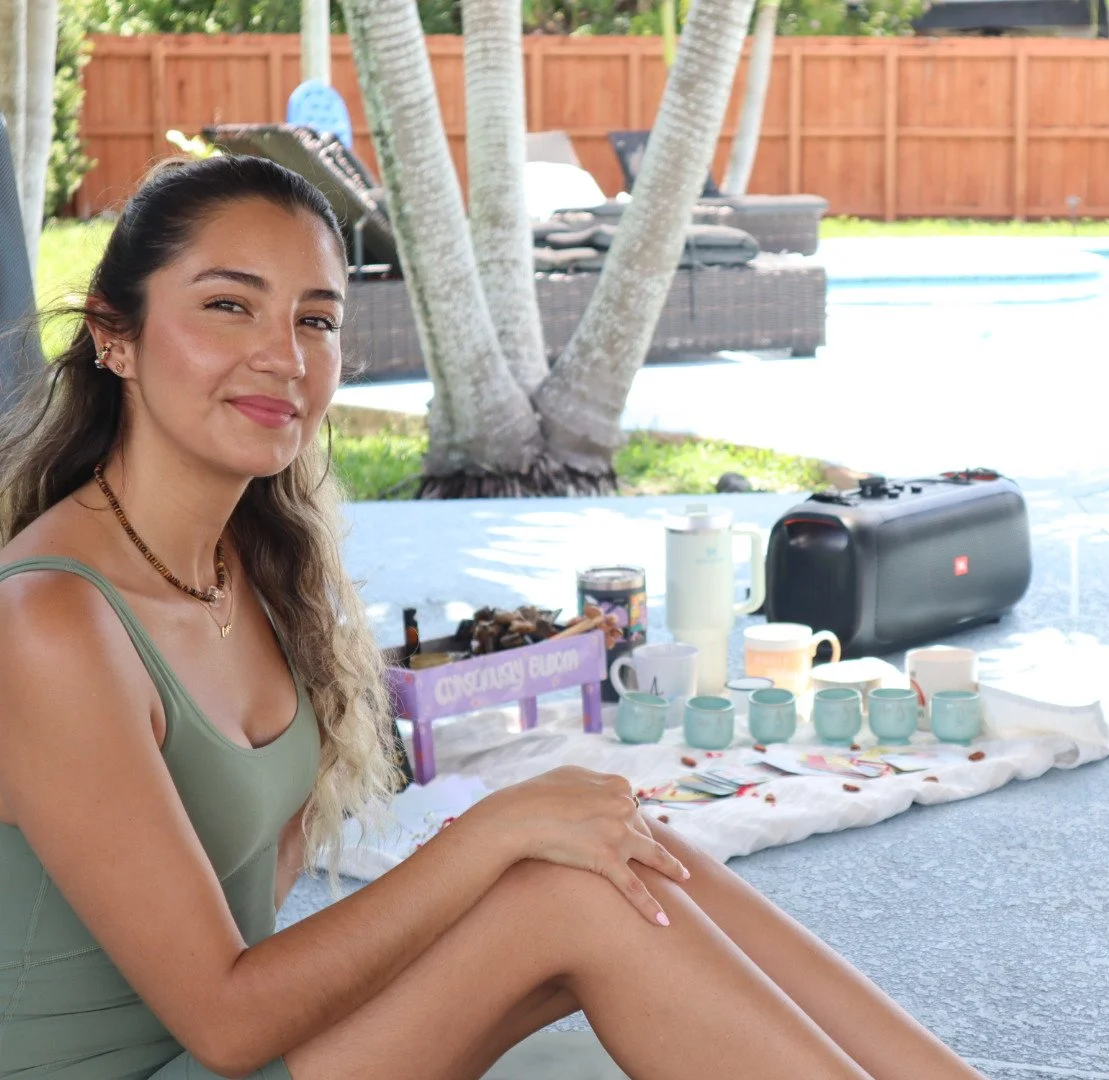 Paola amezquita UKUMANTA Miami  sitting outdoors at a table with a tea set, smiling at the camera, with trees and a pool in the background.