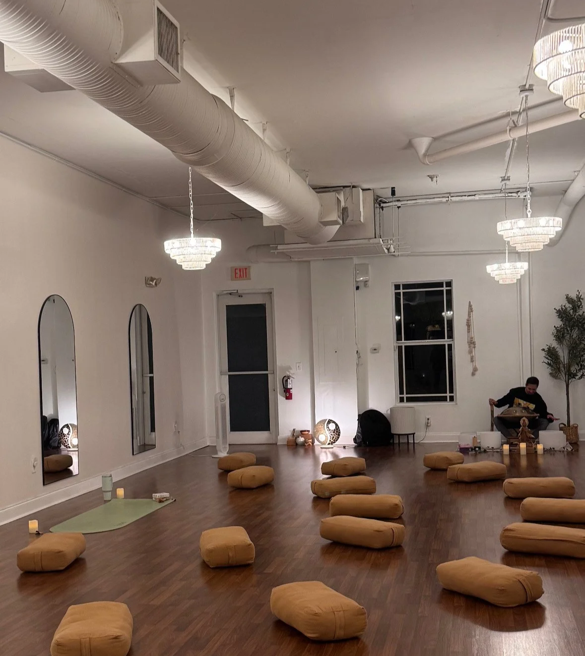 A spacious room with wooden floors and white walls, decorated with hanging chandeliers, a large window, and mirrors. Floor cushions are arranged on the floor, and a person is seated in the background with a musical instrument.