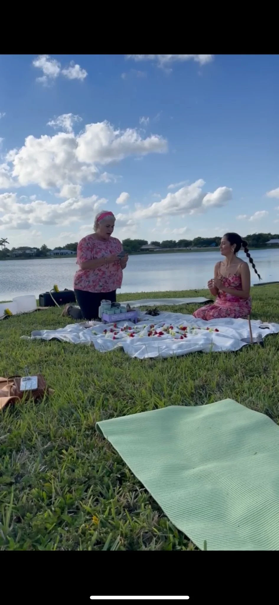 Women having a picnic by a lake under a cloudy sky with a blanket, food, and drinks