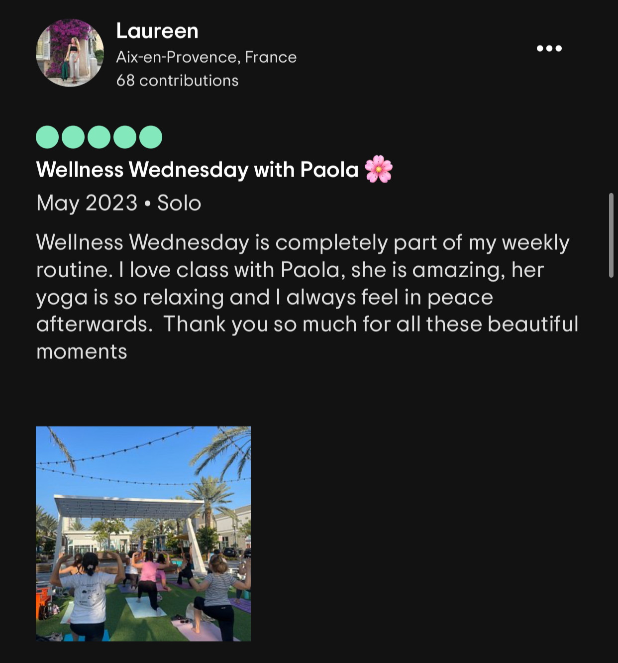Group of people practicing yoga outdoors on mats under a structure with string lights and palm trees during the day.