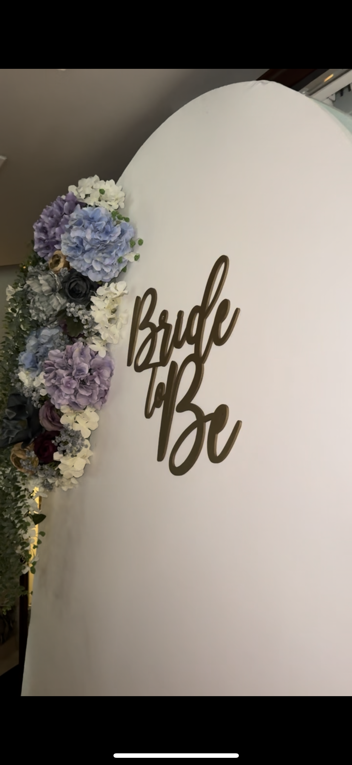 A pregnant woman holding her belly with her left hand, standing in a flower field with pink flowers, wearing a white shirt and jeans.