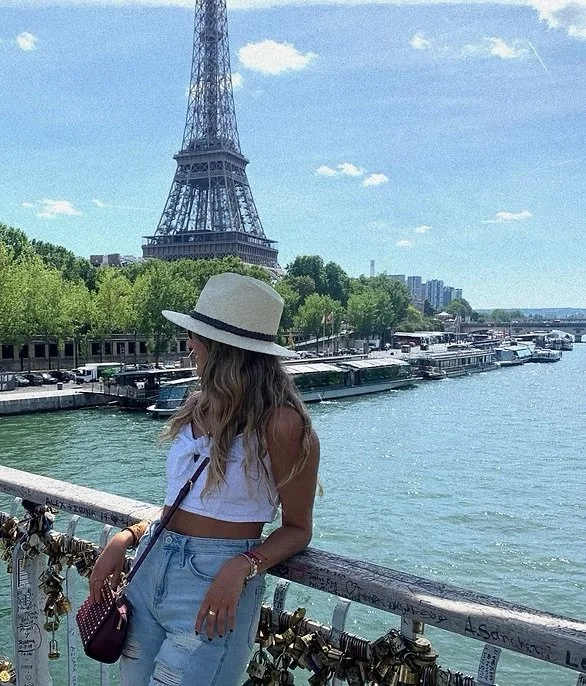 Paola amezquita UKUMANTA Miami  wearing a white sleeveless top, a wide-brimmed hat, and ripped jeans standing on a bridge with padlocks, overlooking the Seine River and the Eiffel Tower in Paris on a sunny day.