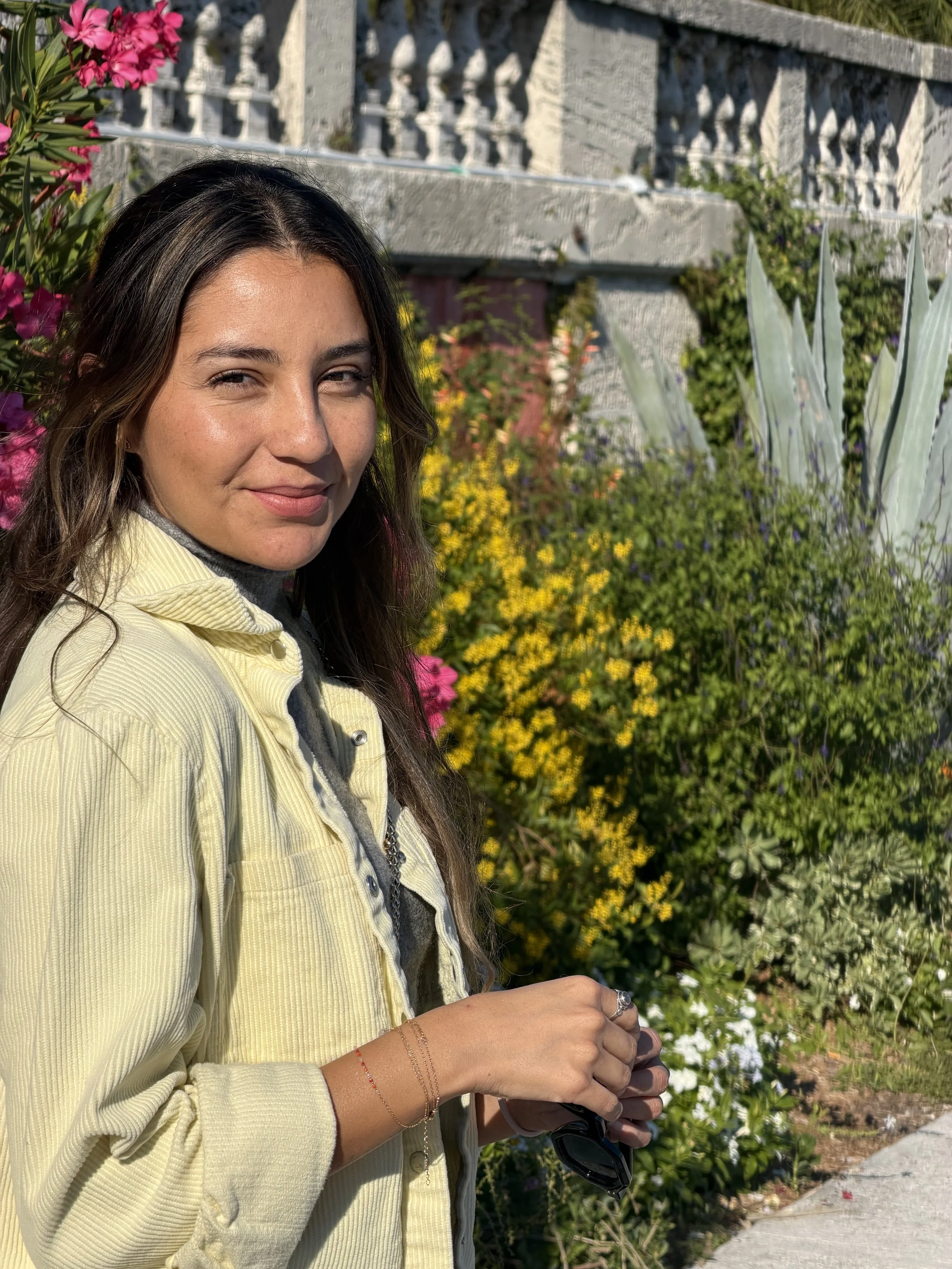 Paola Amezquita with long brown hair standing outdoors in a garden with colorful flowers and succulents, wearing a light yellow jacket and grey shirt, holding sunglasses.