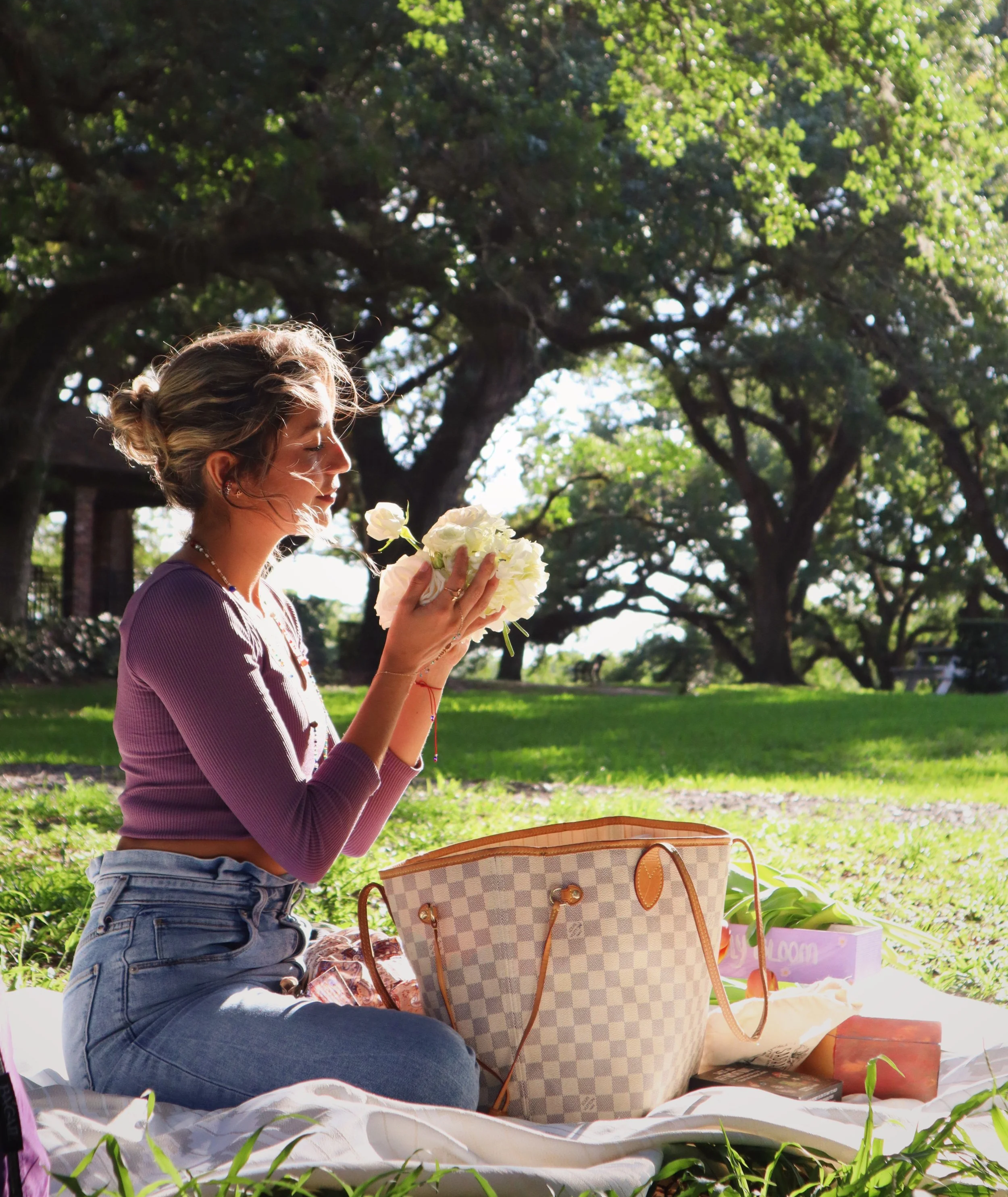 Paola Amezquita sitting on a blanket in a park, holding and smelling a bouquet of white flowers, with a large checkered purse and other items around her, surrounded by lush green trees.