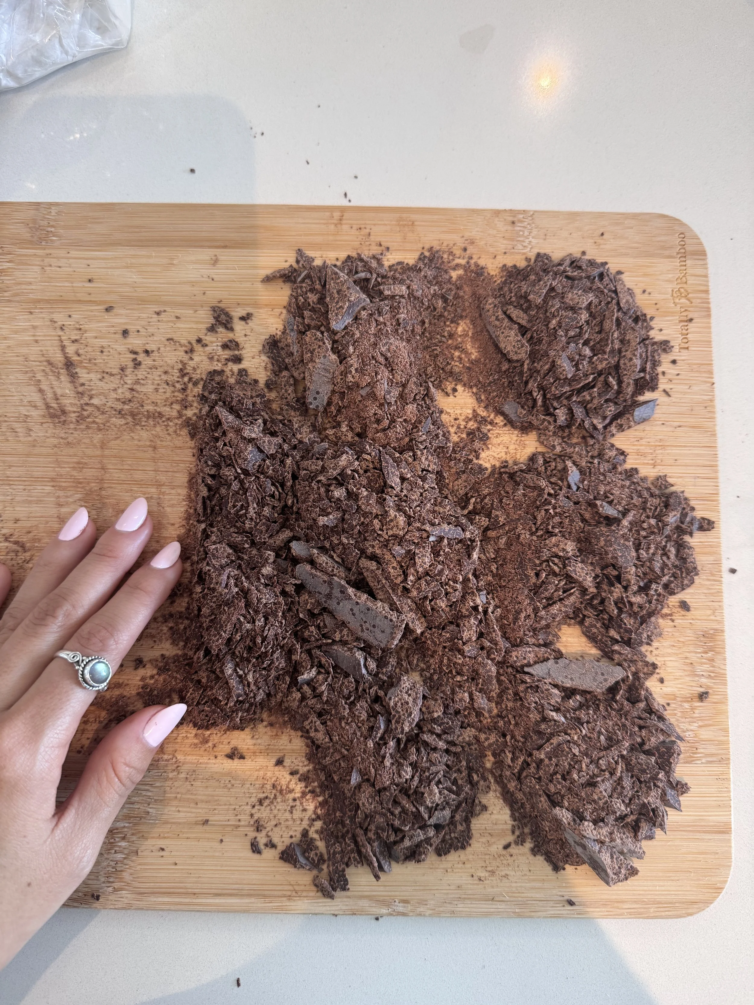 Chopped dark chocolate on a wooden cutting board with a person's hand and ring visible in the lower left corner.