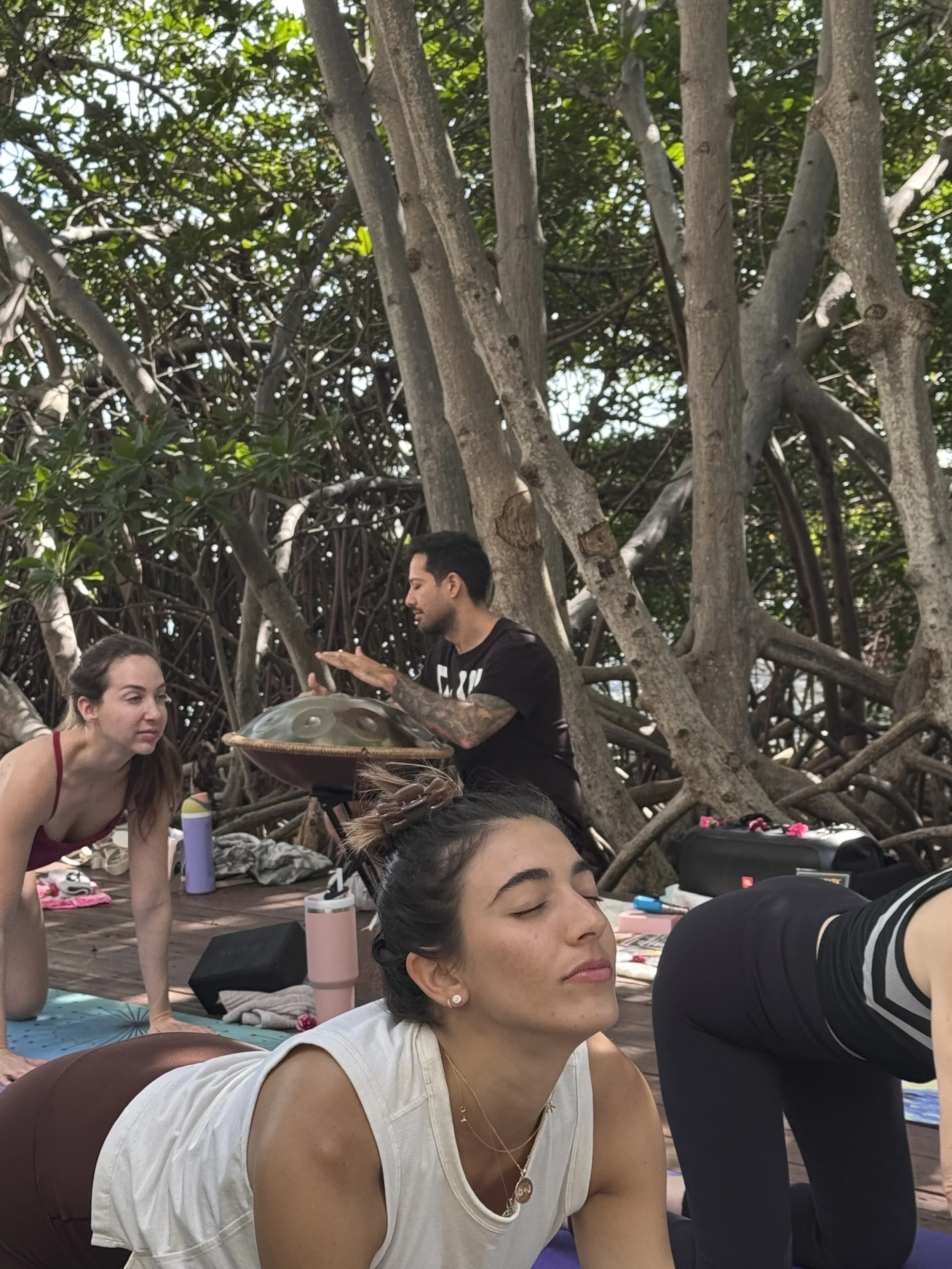 A group of people practicing yoga outdoors under a large tree. The woman in the foreground has her eyes closed, wearing a white top and gold jewelry. In the background, a woman is in a yoga pose, and a man appears to be playing a musical instrument, all surrounded by natural greenery.