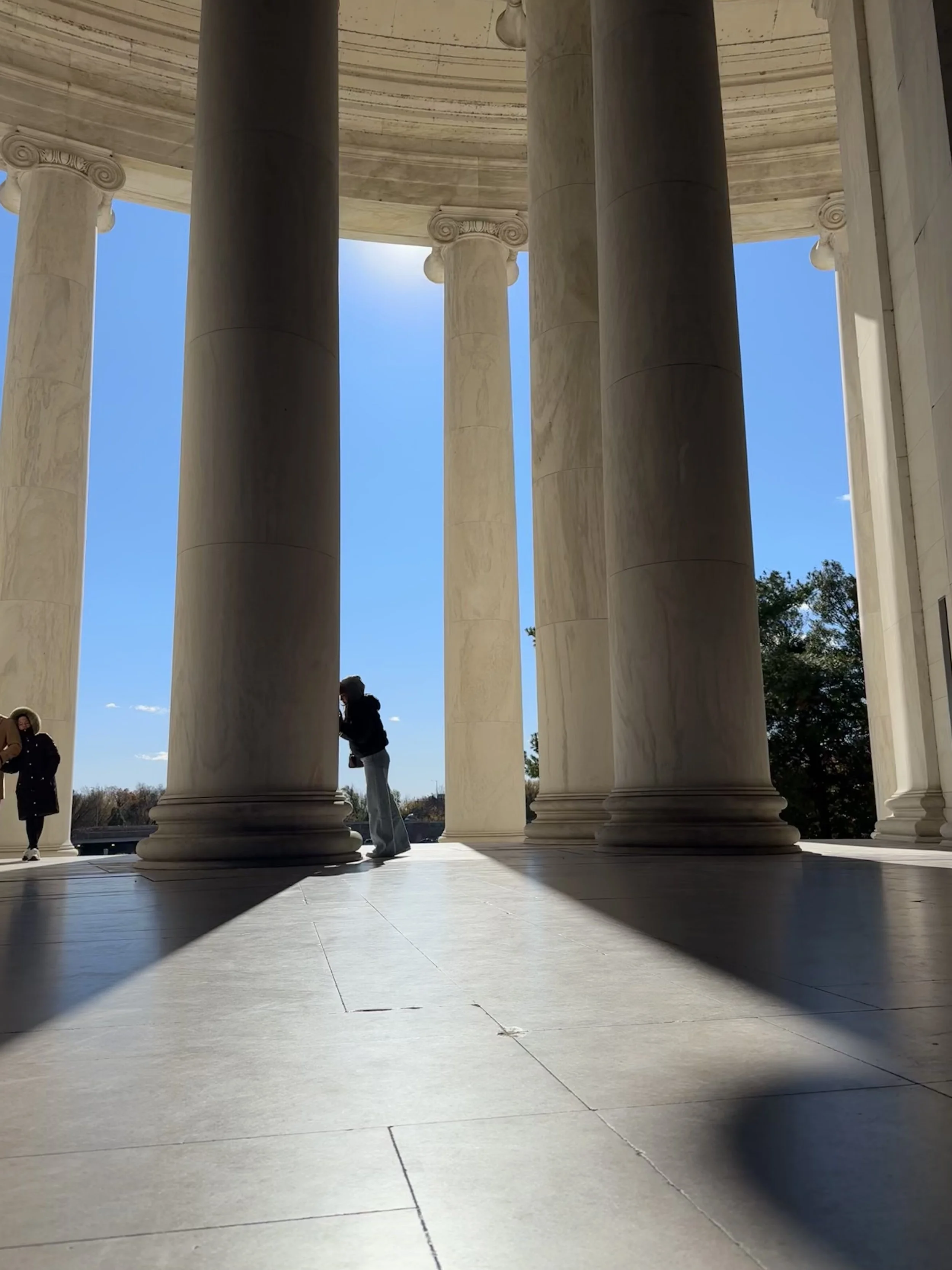 Columned structure with large marble columns casting shadows on the floor, with a clear blue sky in the background and a person standing near a column.