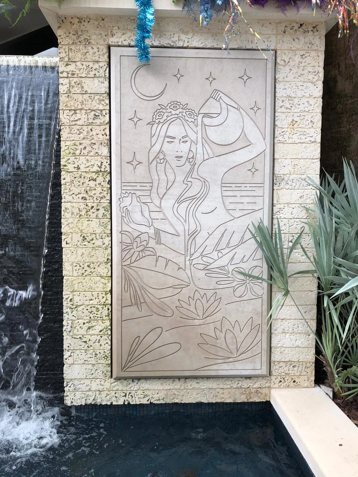 Wall art depicting a woman with long hair, a flower crown, and earrings. She is holding a bottle above her head, pouring liquid into her mouth. The background includes a crescent moon, stars, water, and plants with lotus flowers at her feet.