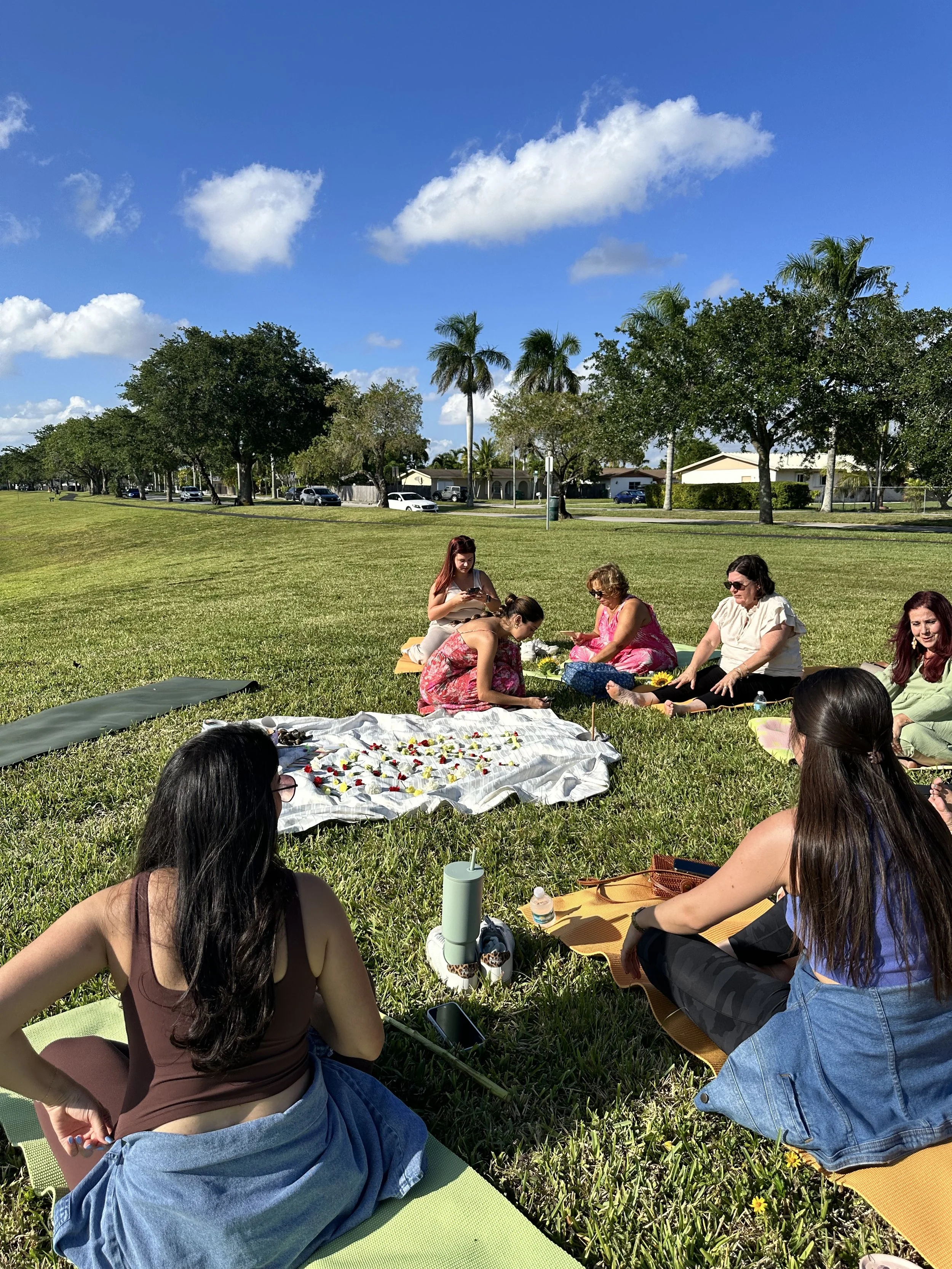 A woman lying on a blanket on the grass with flowers on her chest, a person sitting nearby, and other blankets and belongings in the background.