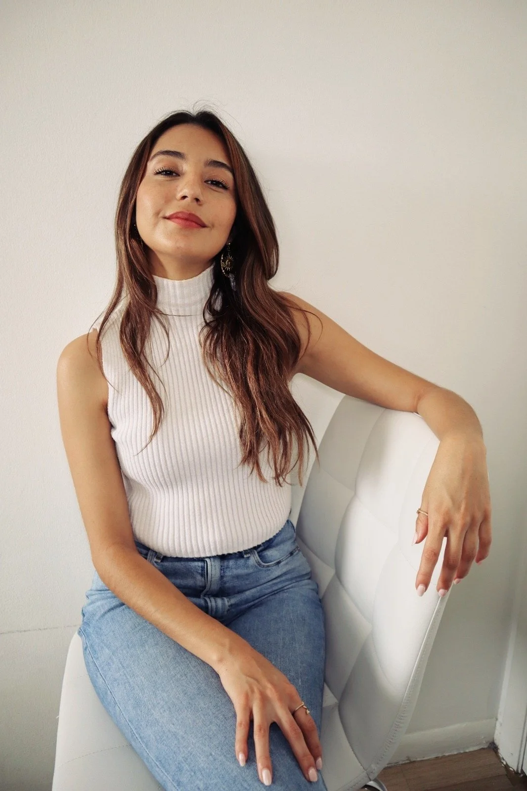 A young woman with long brown hair, wearing a white sleeveless turtleneck top and blue jeans, sitting on a white chair against a plain white wall, smiling slightly with a confident expression.
