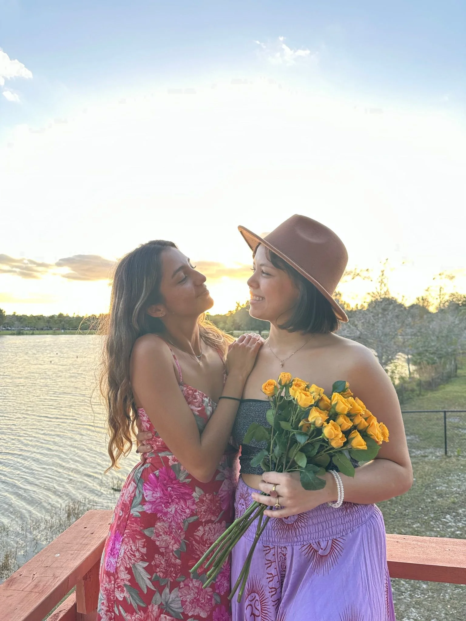 Two women standing on a wooden dock by a lake during sunset, one in a floral dress and the other in a tank top and purple skirt holding a bouquet of yellow roses, smiling at each other.