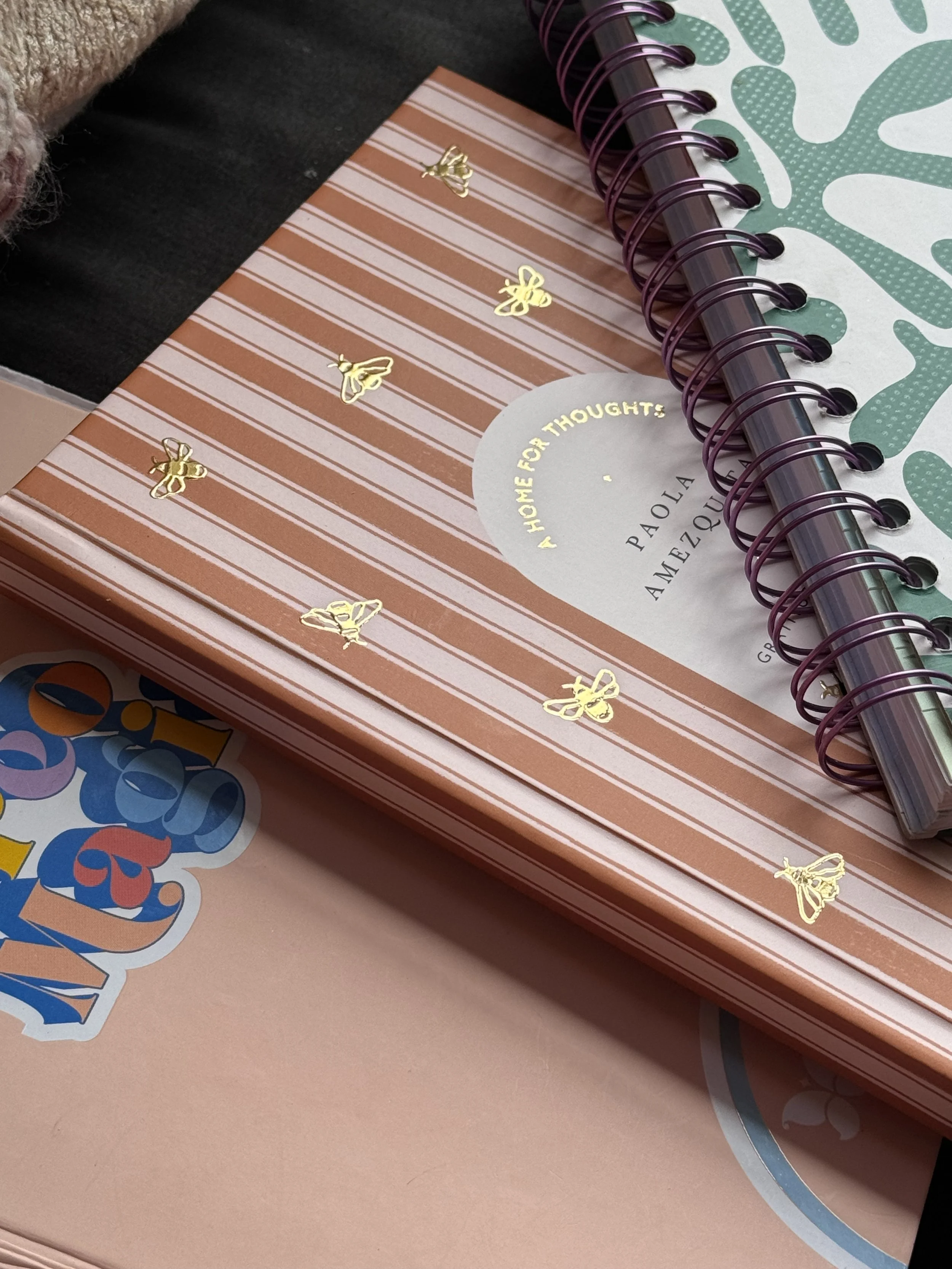 A stack of notebooks and planners, including one with pink and white striped cover decorated with small gold bee illustrations, and another with green and white abstract pattern cover.