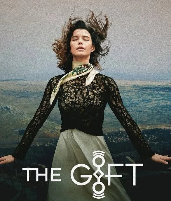 A woman with curly hair standing outdoors against a landscape, wearing a black lace top, a light-colored skirt, and a patterned scarf around her neck. The text "THE GIFT" is overlaid at the bottom of the image.