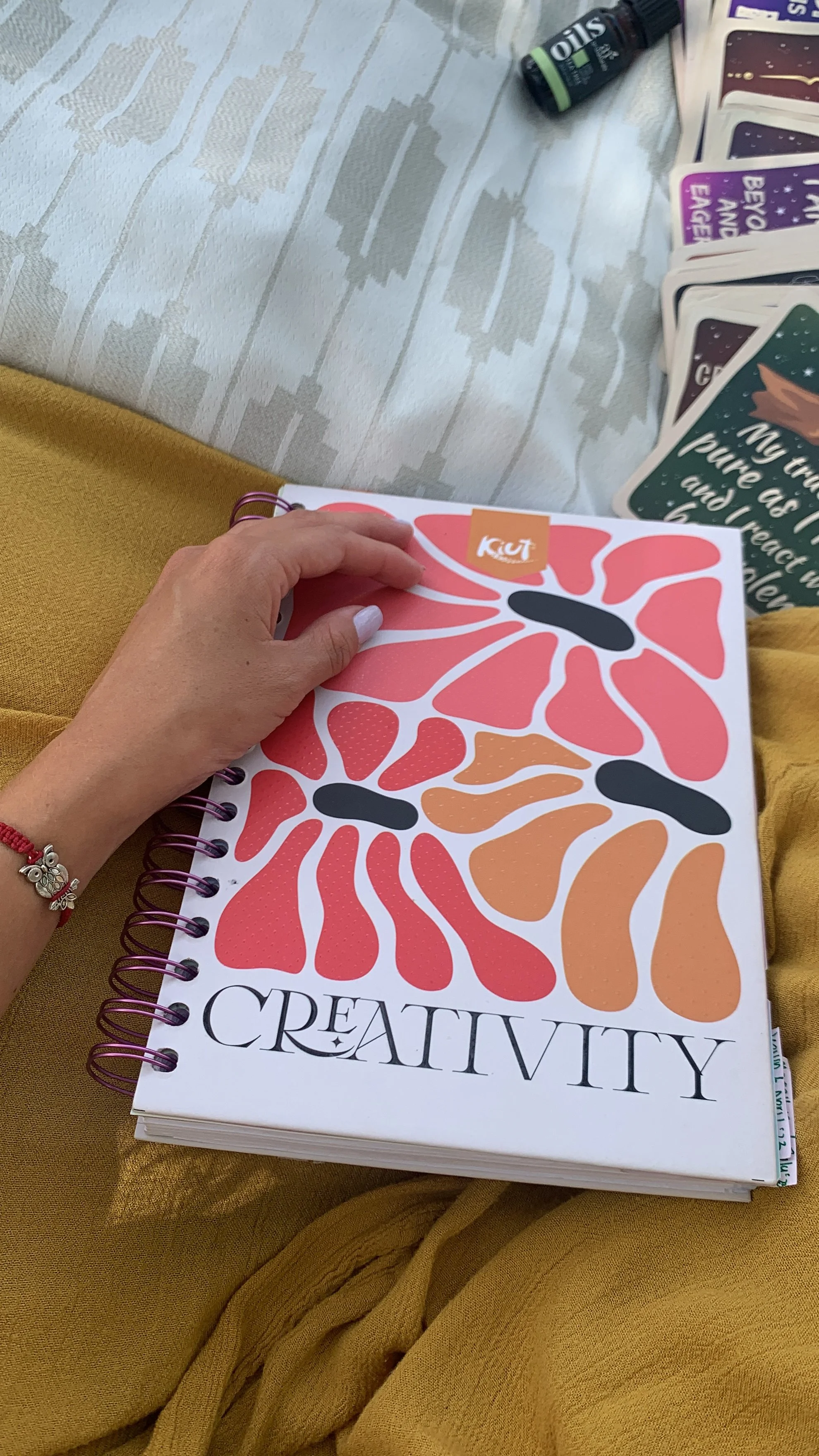 A hand holding a notebook with a colorful floral design labeled 'CREATIVITY', with various cards partially visible in the background and art supplies on a table.