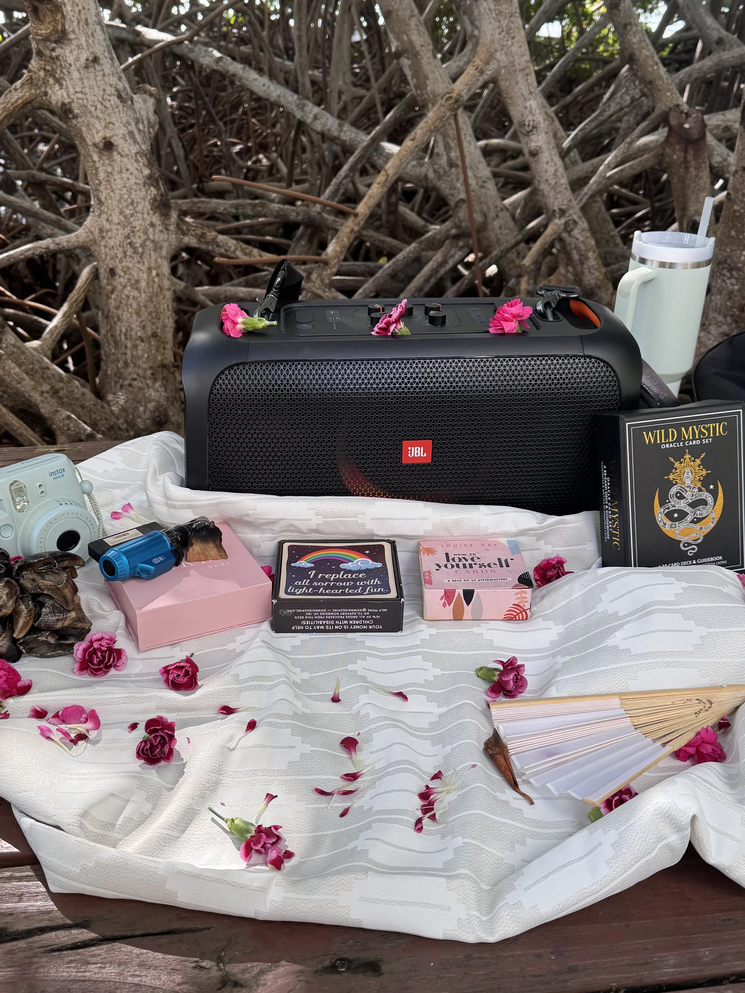 A table set outdoors with objects including a JBL speaker with pink flowers, tarot cards, a fan, a small blue spray bottle, a camera, a white tumbler with a straw, a black box labeled 'Wild Mystic' with a snake illustration, and a decorative fan, all on a white cloth with pink flower petals scattered around, against a background of tree branches.