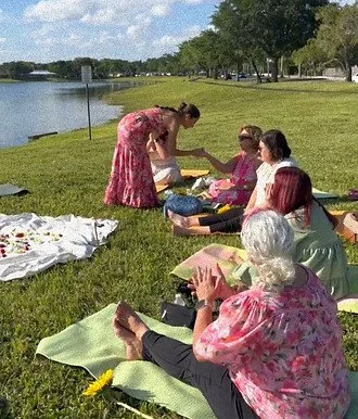 Group of women sitting on blankets by a lake, enjoying a picnic, with one woman handing food to another.