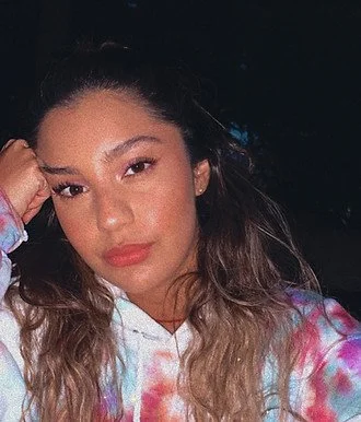 A young woman with long, wavy hair and a colorful tie-dye hoodie, posing with her hand resting on her head against a dark background.