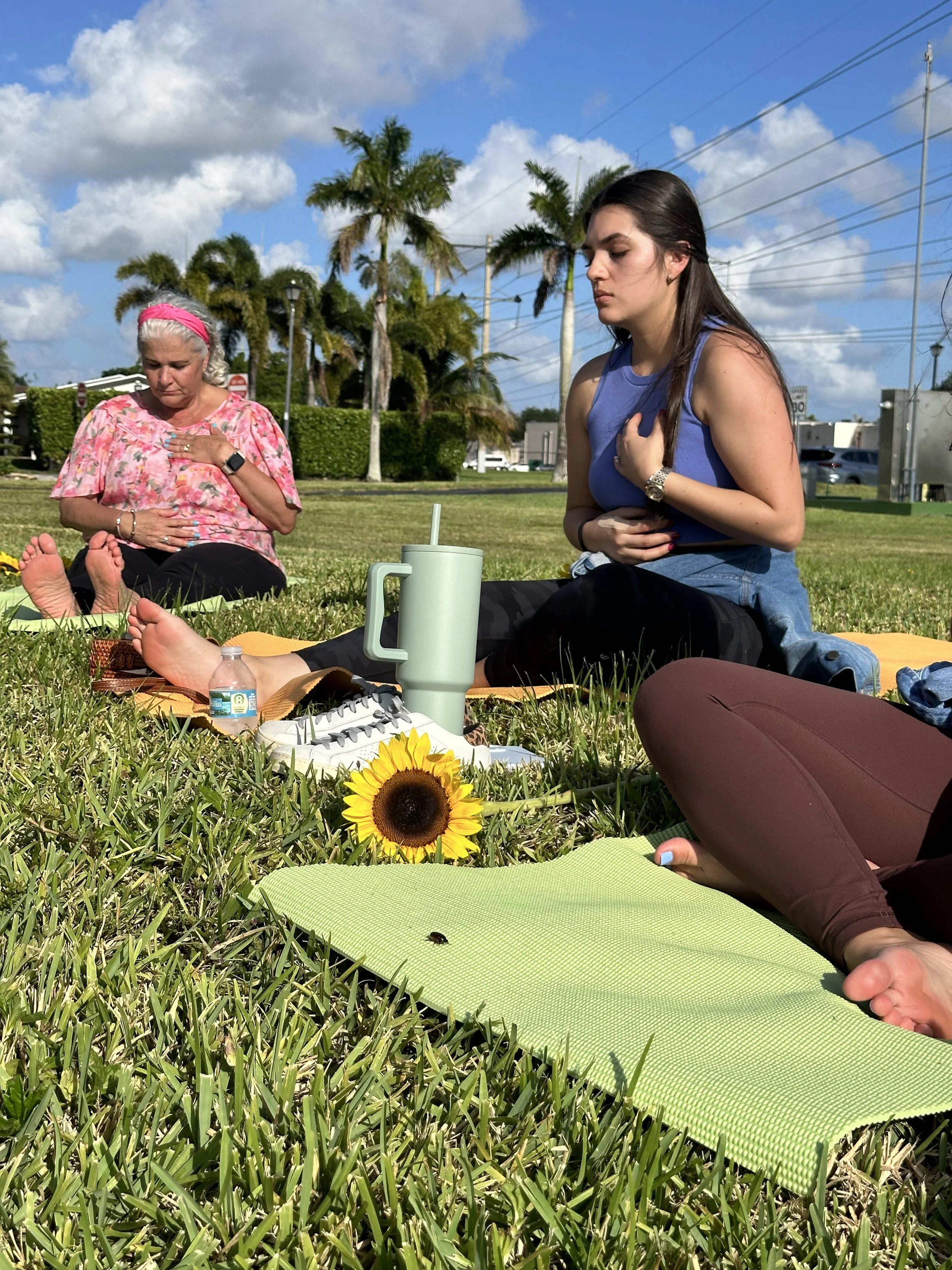 Three women sitting on the grass with eyes closed and hands over their chests, participating in a meditation or mindfulness session outdoors. There are yoga mats, a sunflower, a water bottle, and a cup with a straw on the grass, with palm trees and a blue sky with clouds in the background.