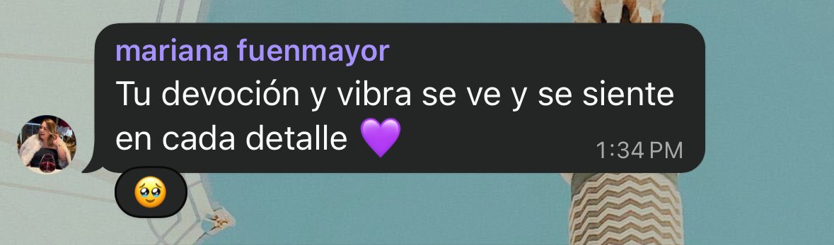A text message in Spanish from Mariana Fuenmayor saying: "Tu devoción y vibra se ve y se siente en cada detalle" with a purple heart emoji, sent at 1:34 PM, on a chat overlayed on an outdoor photo with a person wearing a zigzag patterned dress and a 