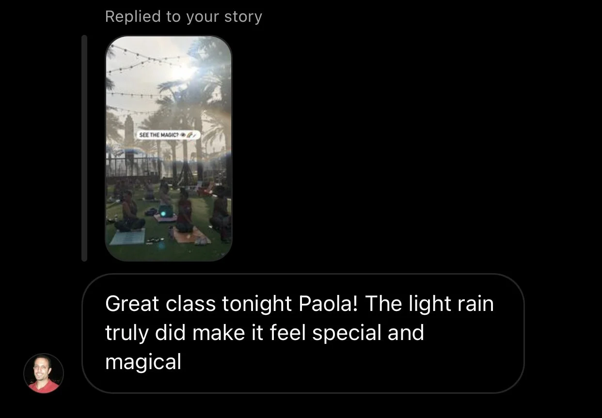 Group of people sitting in a circle on the grass outdoors, with palm trees and string lights in the background, participating in a meditation class during light rain.