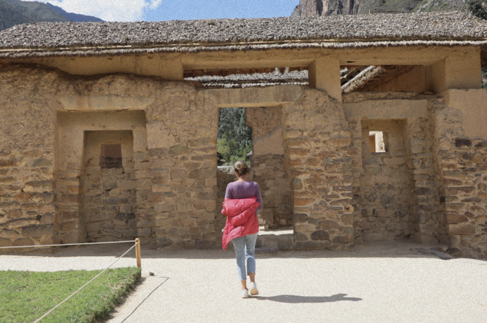 Woman walking towards ancient stone structure with rectangular doorways, greenery in background, sunny day.