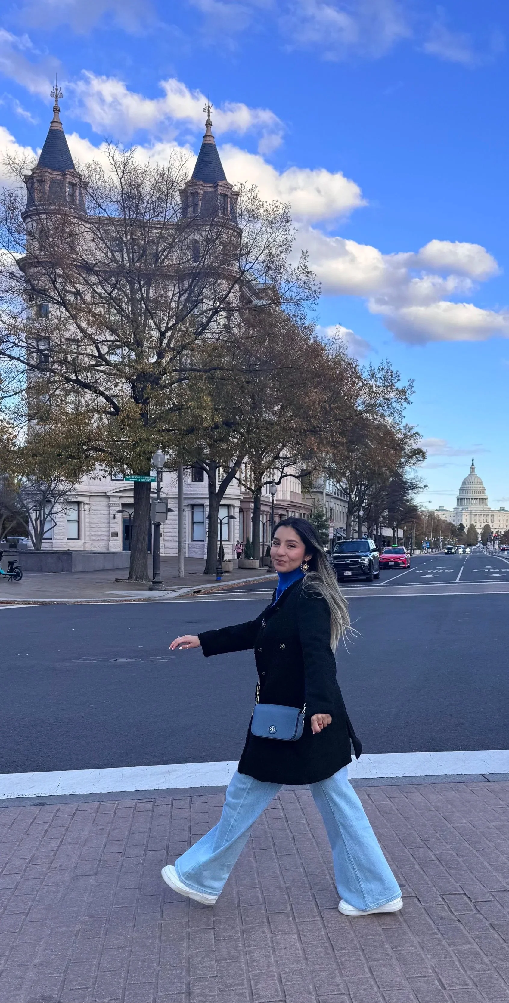 A woman in a black coat and light blue wide-leg jeans crossing a street in front of the U.S. Capitol building with a cloudy sky above.