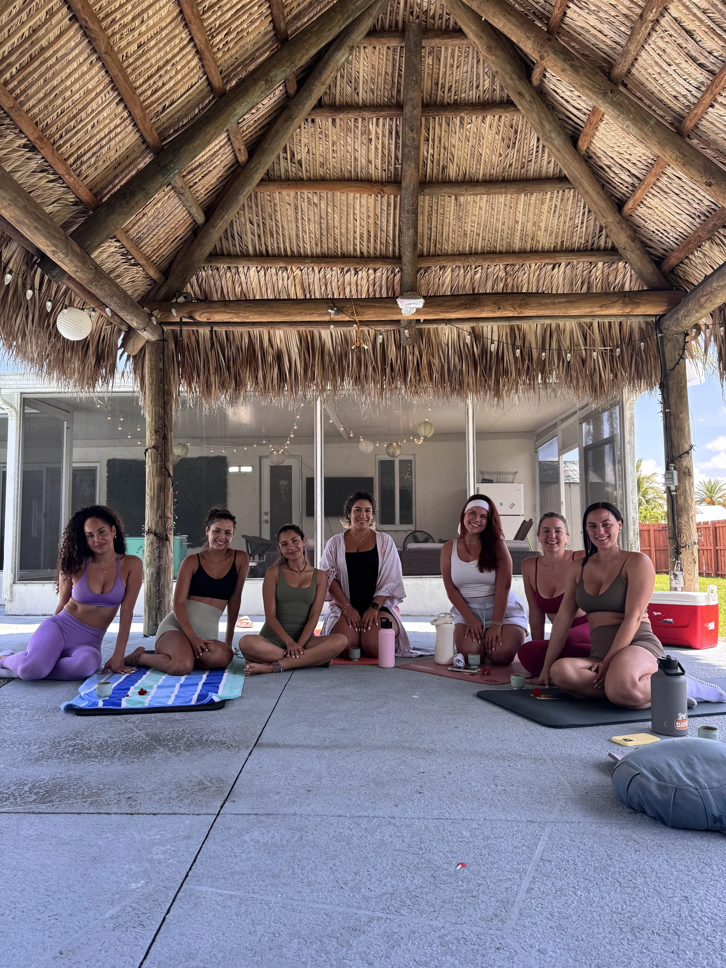 Group of seven women sitting cross-legged under a thatched roof, participating in a yoga session with yoga mats, water bottles, and candles, in an outdoor patio area.