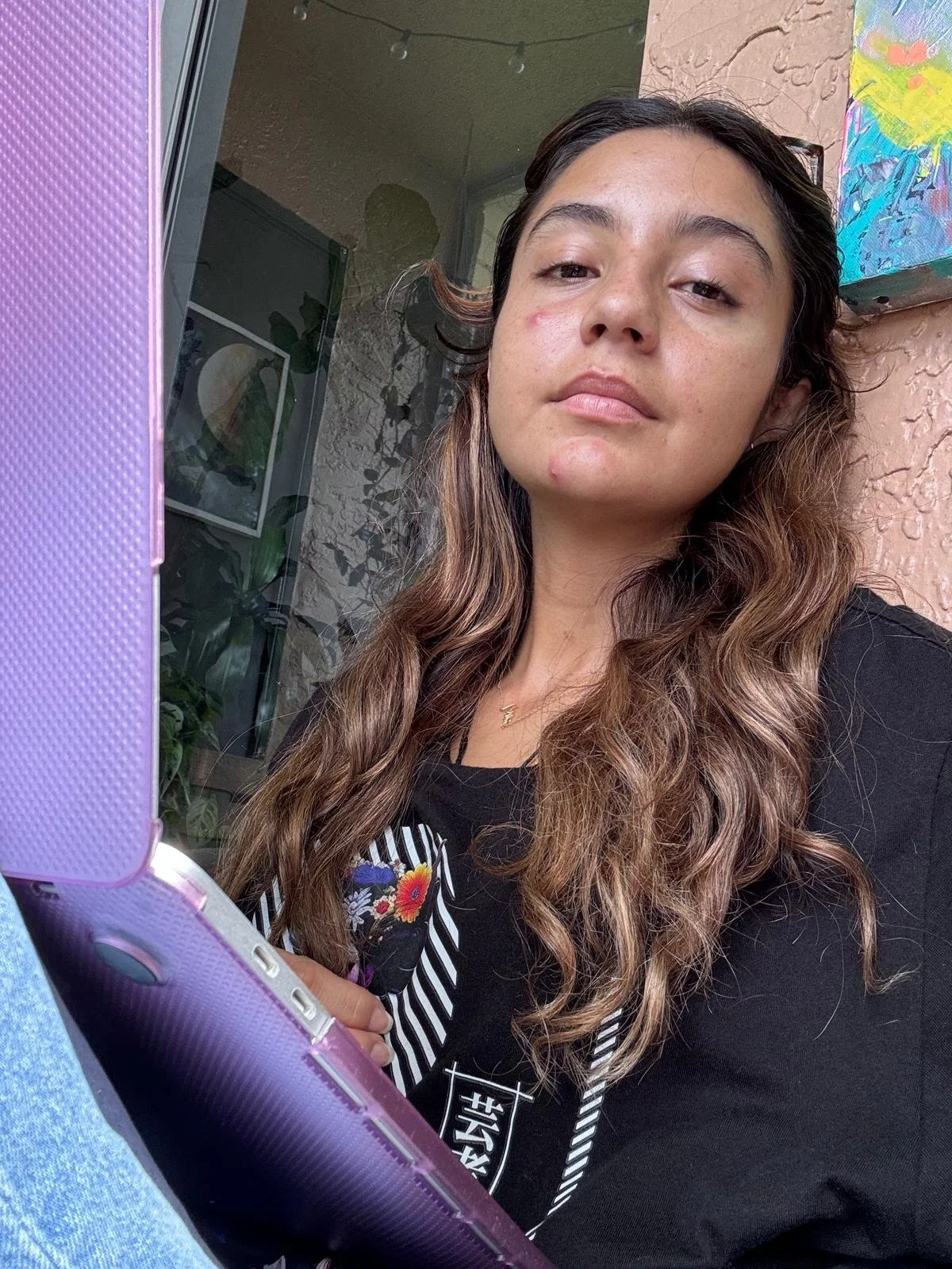 A young woman with wavy brown hair holding a pink phone with a purple case, sitting outdoors near a glass door and pink stucco wall, looking at the camera with a neutral expression.