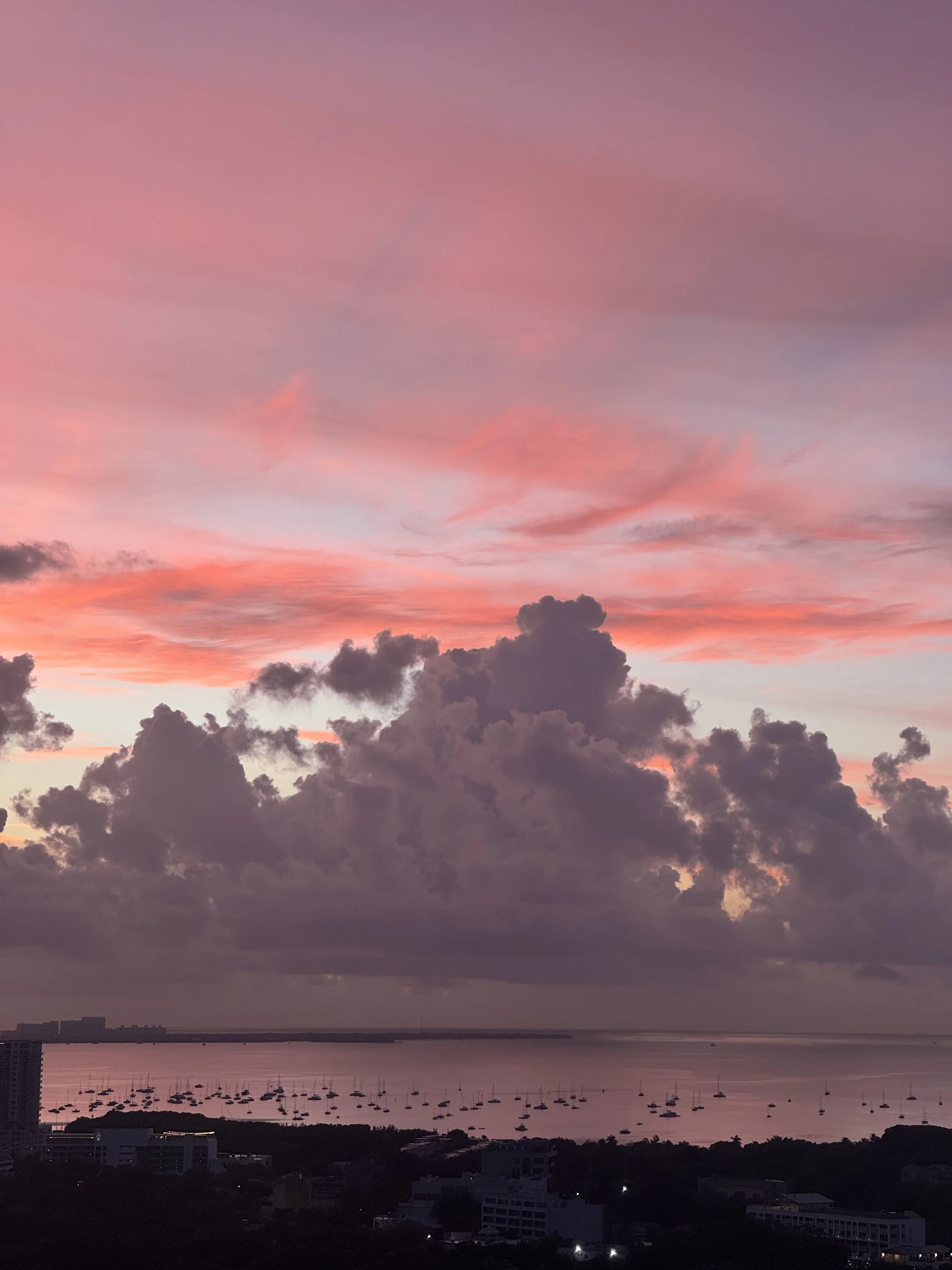 A sunset over a coastal city with pink and purple clouds and a body of water with sailboats.
