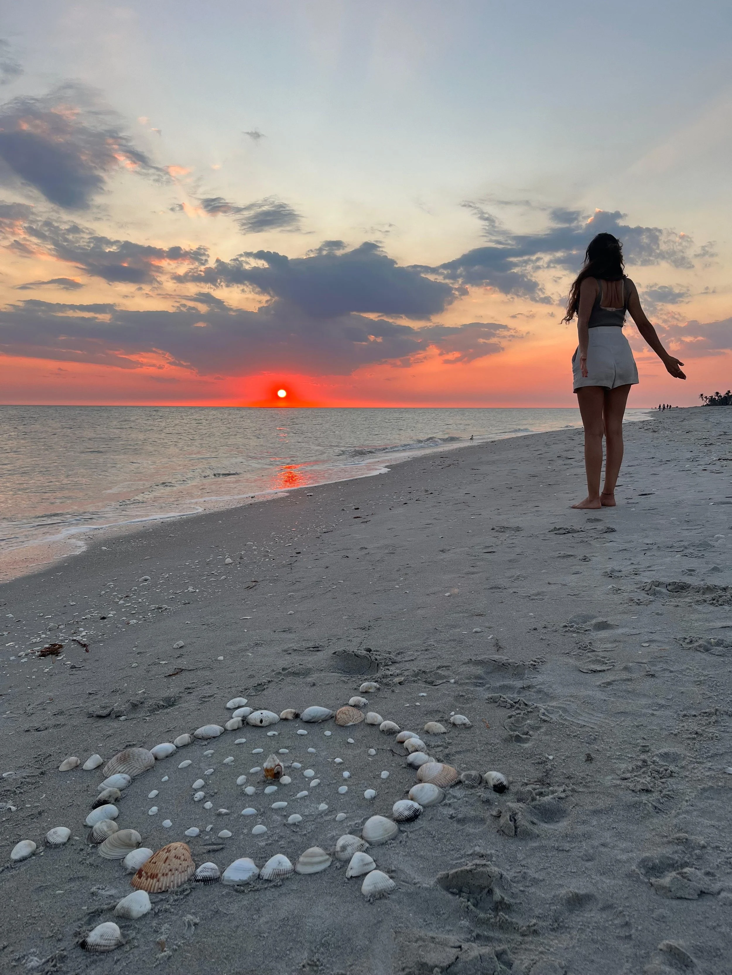 A woman in a black tank top and white shorts standing on a sandy beach at sunset, with a heart-shaped design made of seashells in the sand in the foreground, and the ocean and sky with colorful clouds in the background.