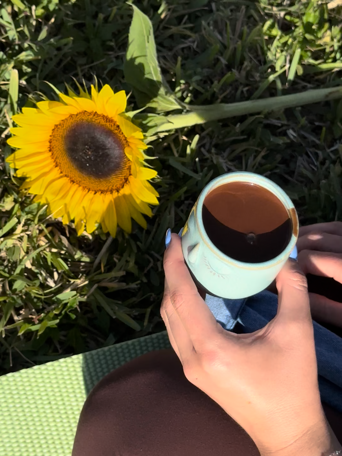 A person holding a mug filled with coffee while sitting outdoors on green grass, with a sunflower next to them and sunlight casting shadows.