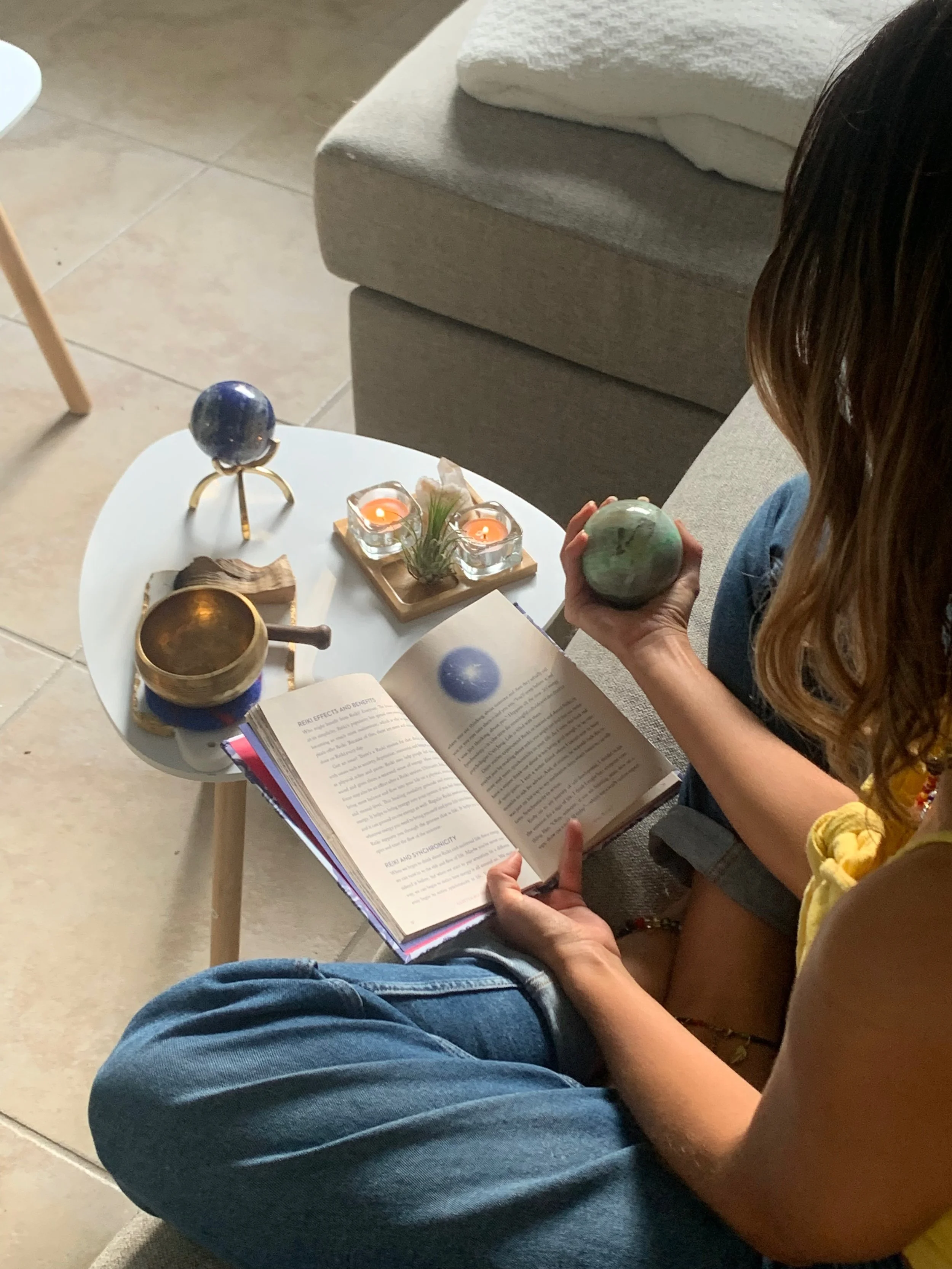 Person sitting cross-legged reading a book, holding a green marble, with a small table beside them featuring mini globes, candles, and a singing bowl.