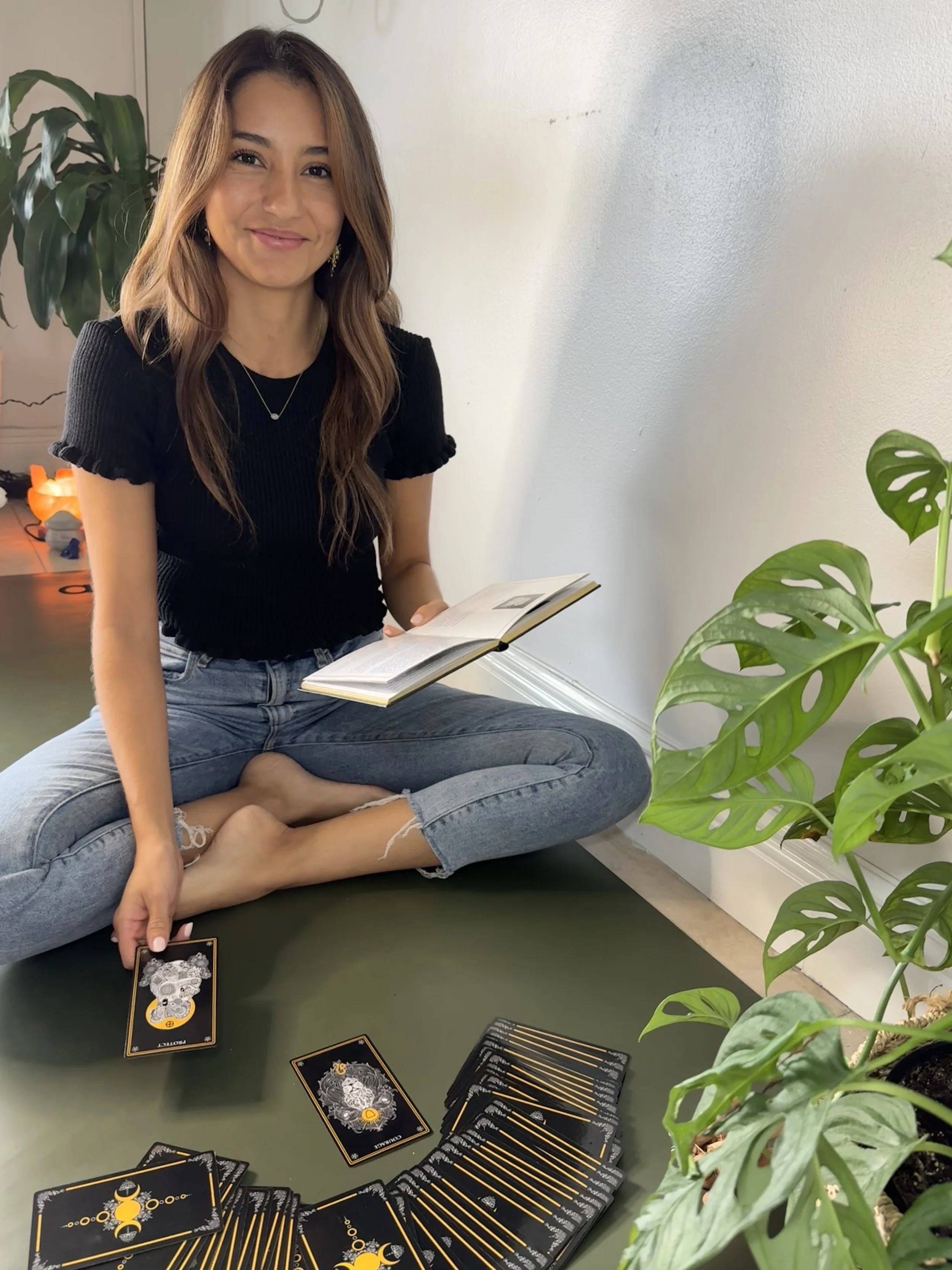 Paola Amezquita sitting cross-legged on the floor, holding tarot cards in one hand and a book in her lap, next to a large green plant.