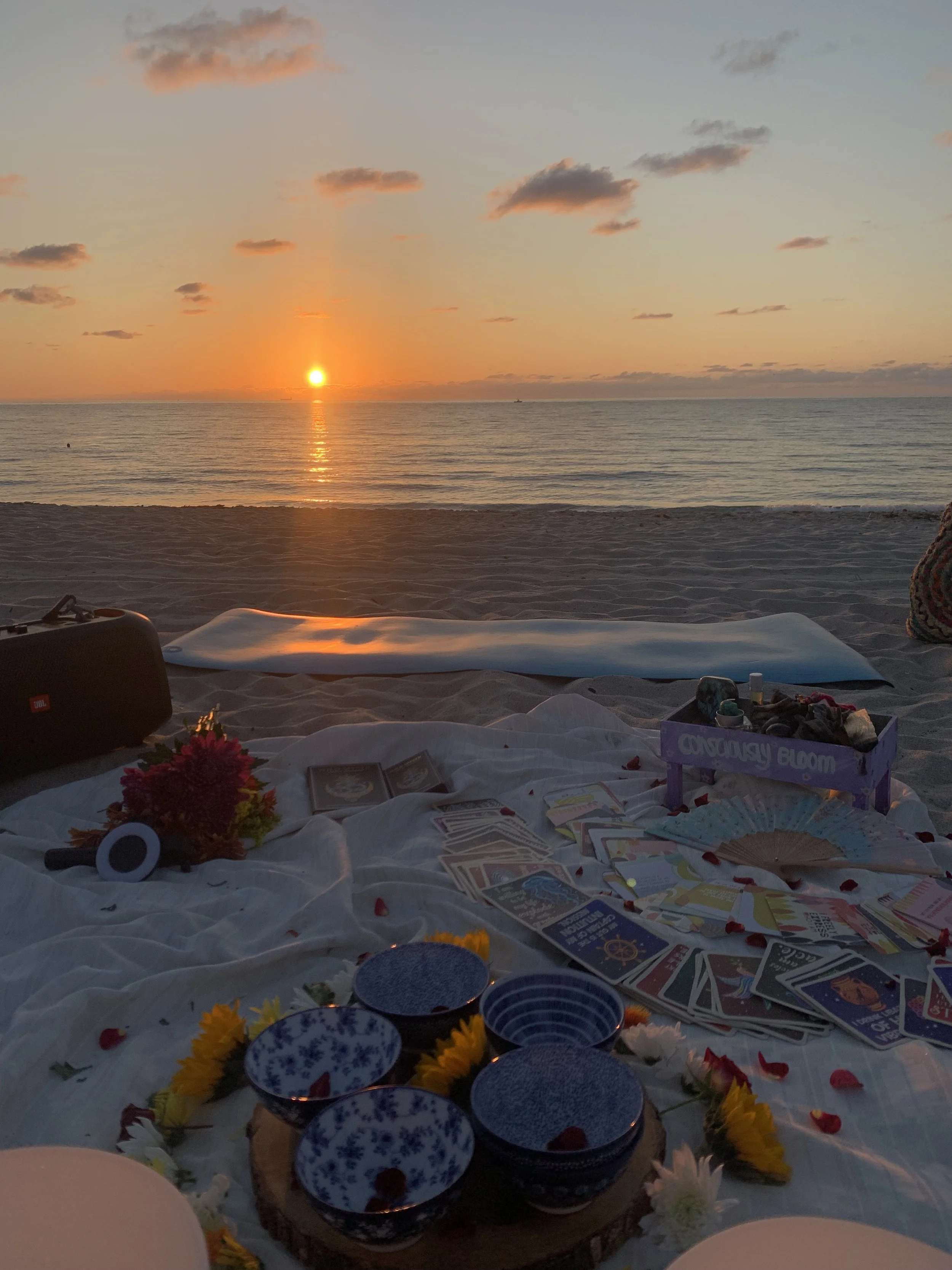 Beach scene at sunset with a picnic setup on the sand, including tarot cards, bowls, flowers, and a speaker, with the ocean and sunset in the background.